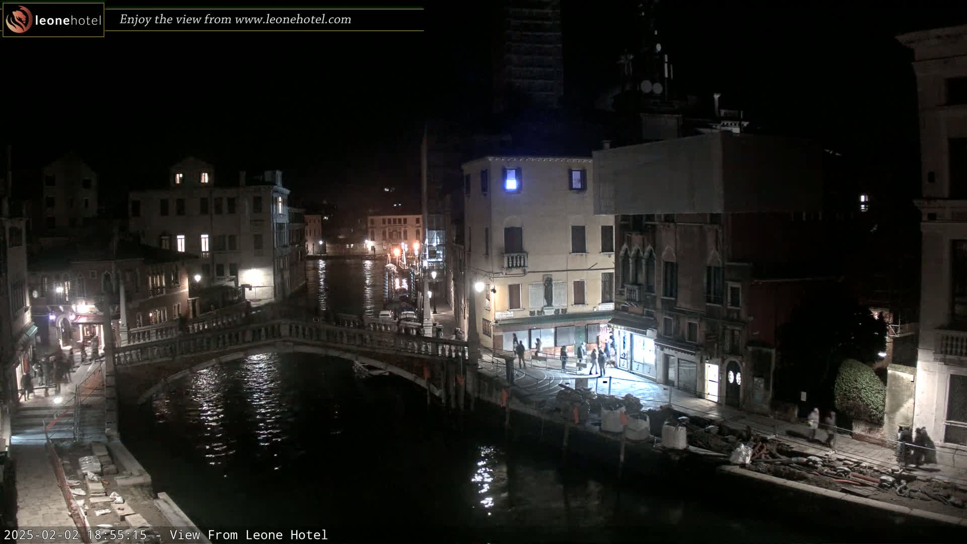 A nighttime view of a canal in Venice, Italy, shows a bridge, several buildings, and some people walking along the water's edge.