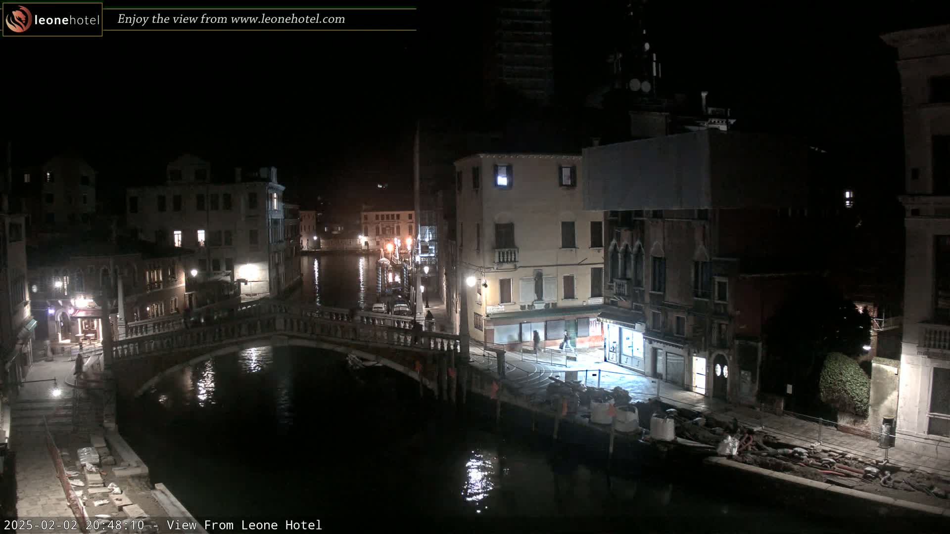 A nighttime view of a canal in Venice, Italy, shows several buildings lining the waterway, with a bridge crossing the canal and some construction materials visible on the bank.