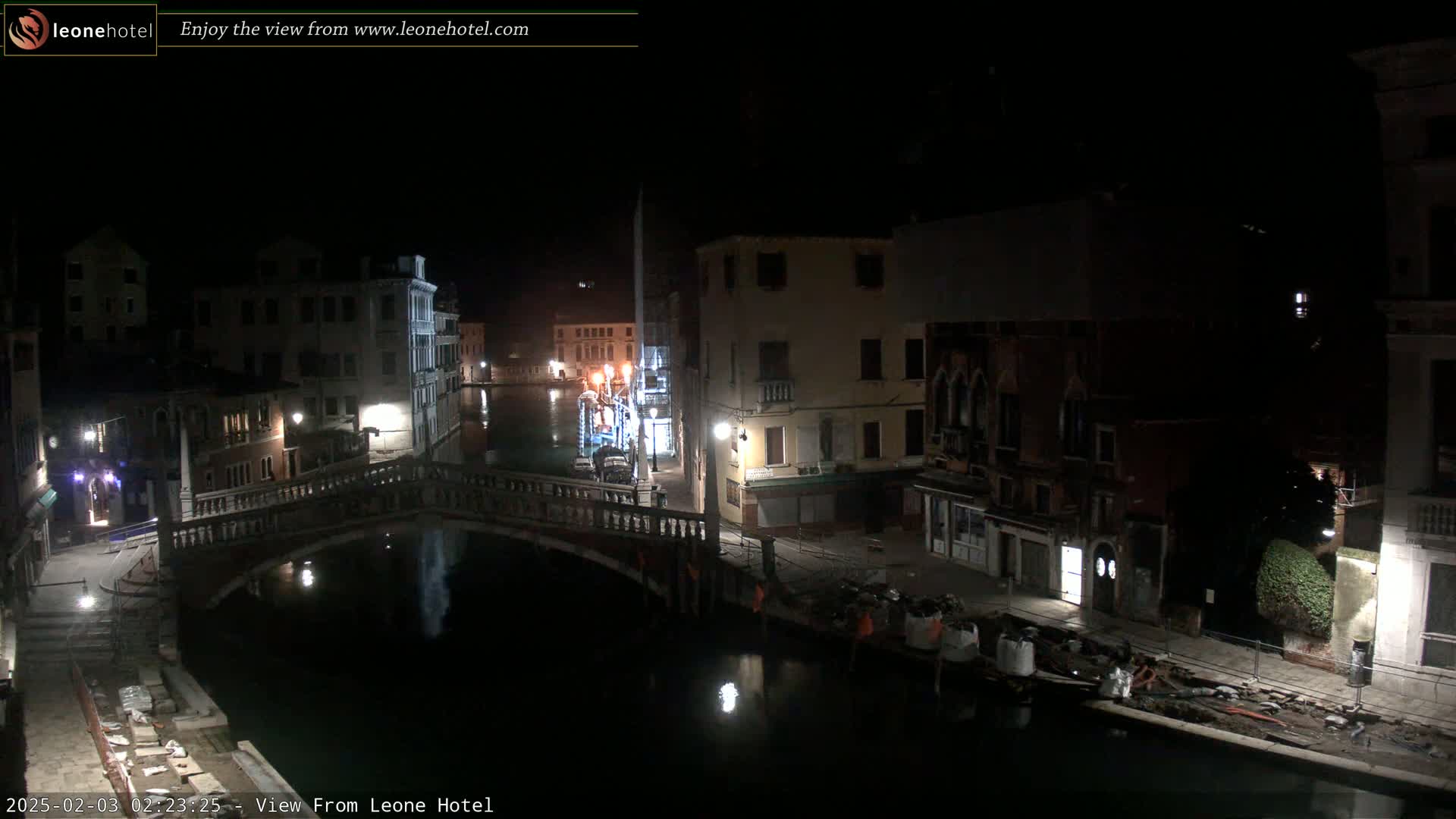 A nighttime view of a canal in Venice, Italy, shows a bridge and buildings along the waterway, with some construction or repairs visible along the canal bank.