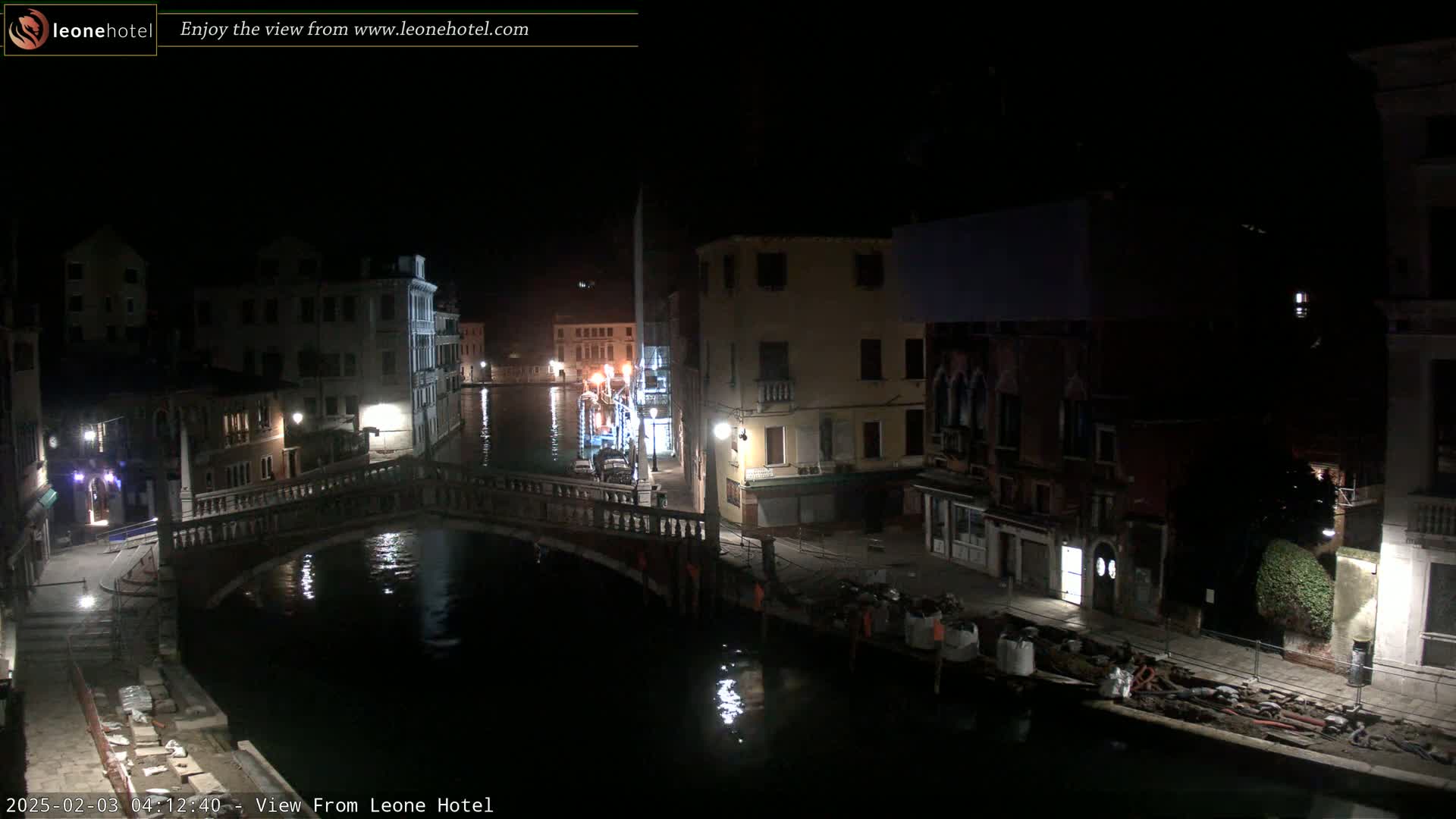 A nighttime view of a canal in Venice, Italy, shows buildings lining the waterway with a bridge crossing the canal, and some construction materials visible near the water's edge.