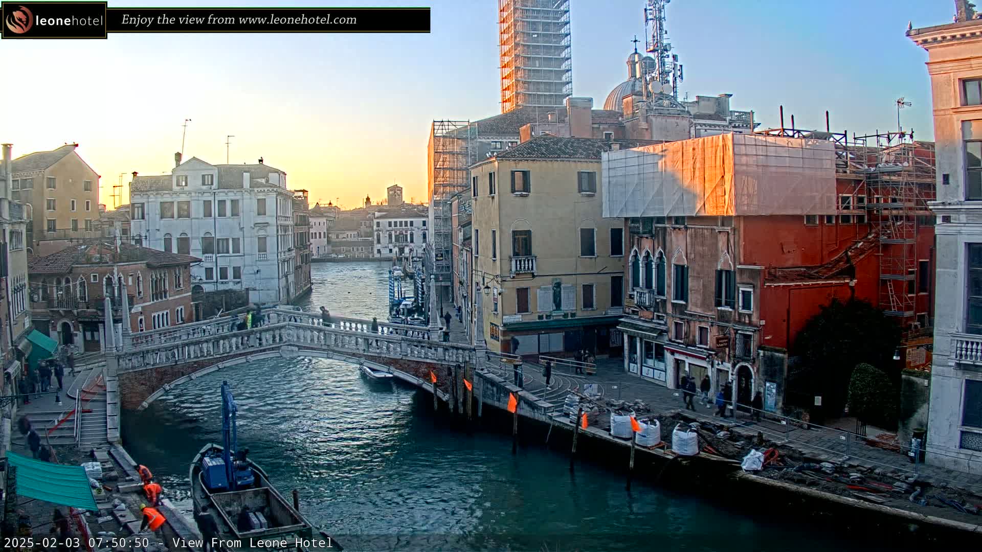 A Venetian canal at sunset, showing a stone bridge, several buildings under construction, and a small boat near the bridge.