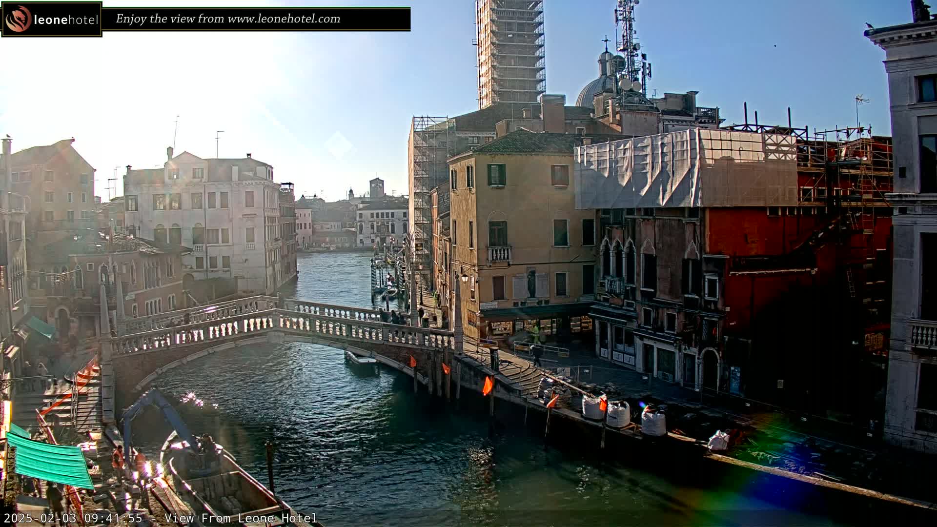 A sunny day in Venice shows a canal with a stone bridge, several buildings, some under construction, and boats moored along the waterway.