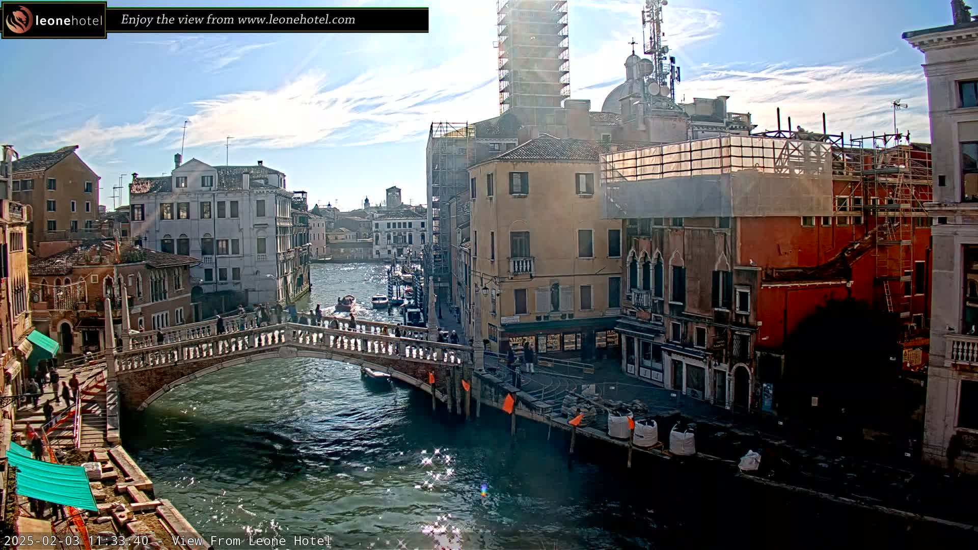 A sunny day in Venice shows a canal with a stone bridge, several buildings undergoing construction or renovation, and boats on the water.