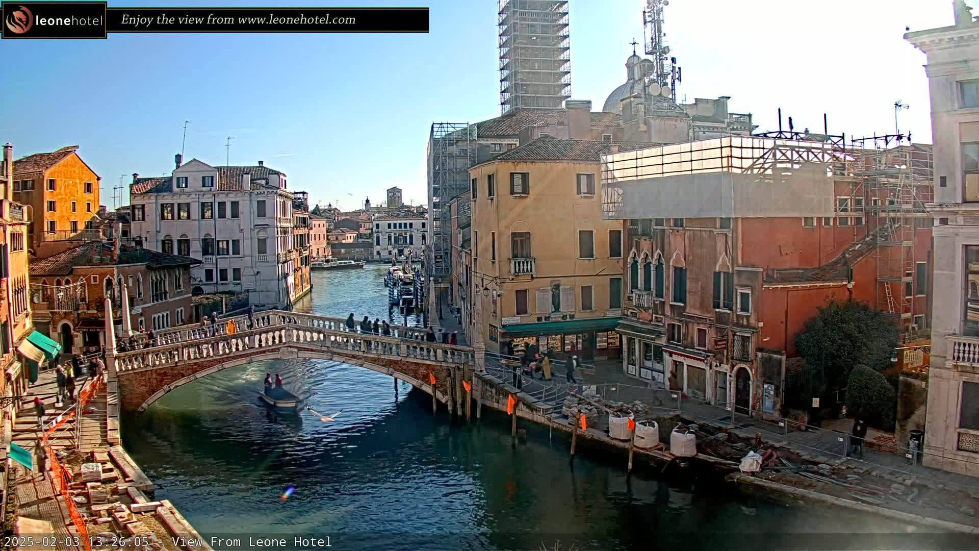 A sunny day in Venice shows a canal with a stone bridge, several buildings undergoing renovations, and a gondola passing underneath the bridge.