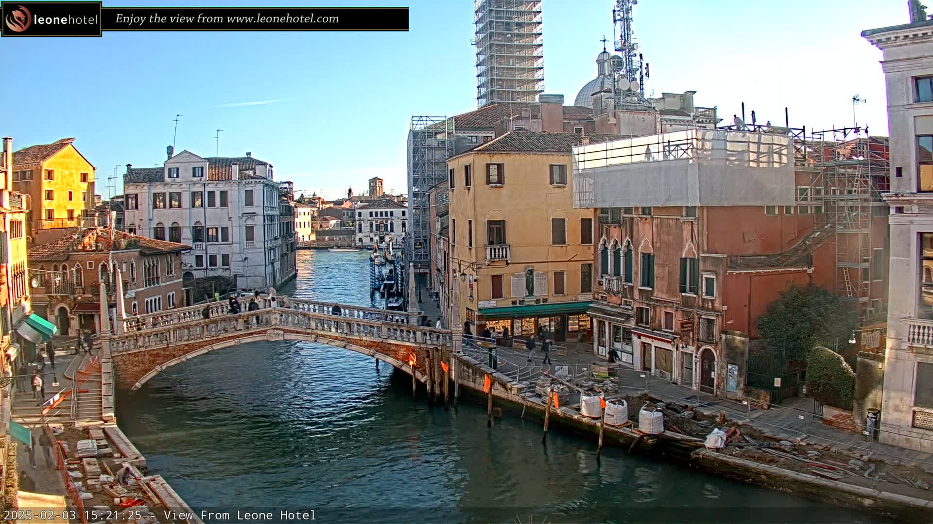 A Venetian canal scene on a sunny day shows a stone bridge arching over the water, with colorful buildings lining the waterway, some under construction, and small boats moored along the banks.