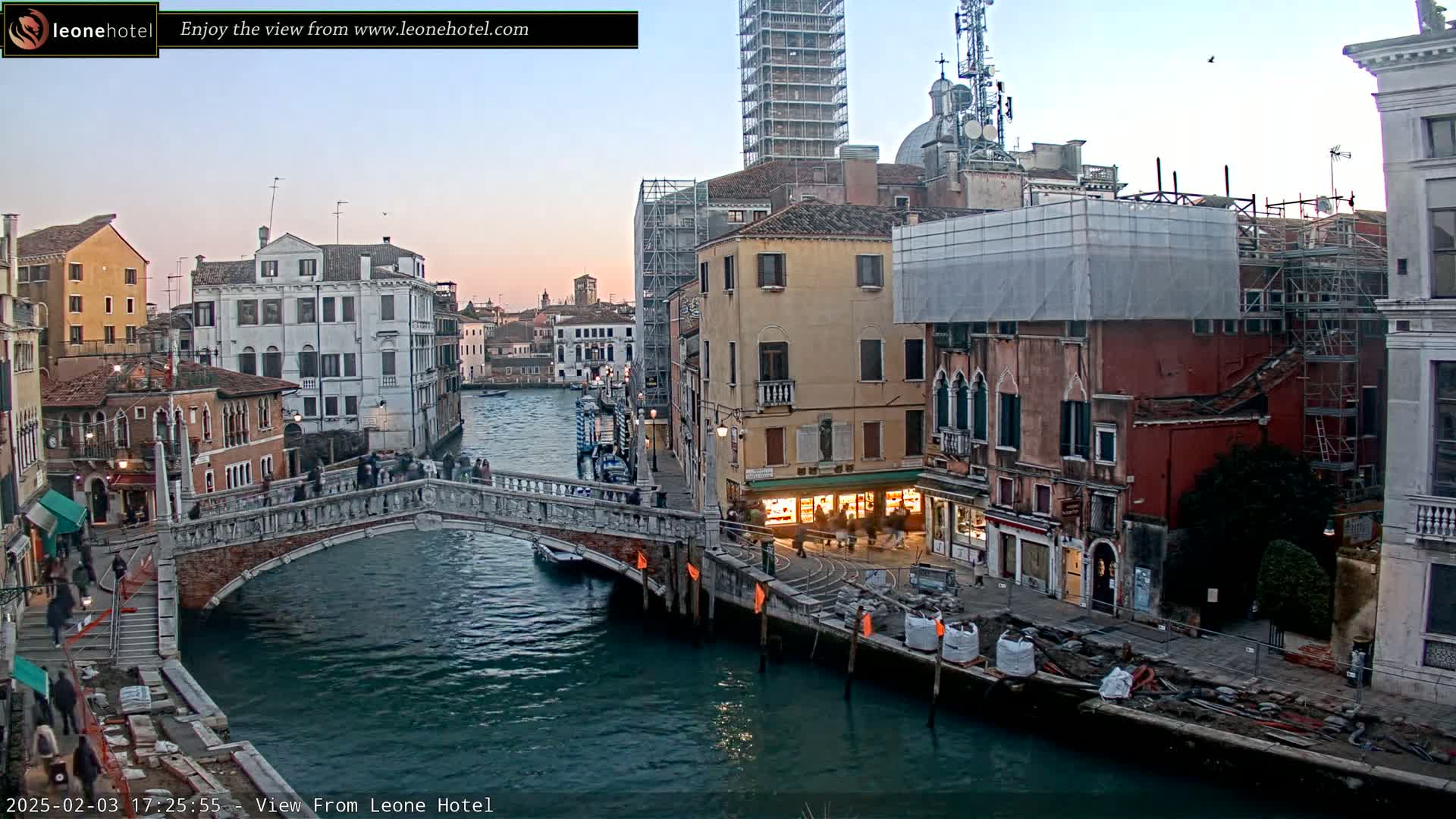 A stone bridge spans a canal in Venice, Italy, at dusk, with several buildings lining the waterway, some undergoing construction or renovation.