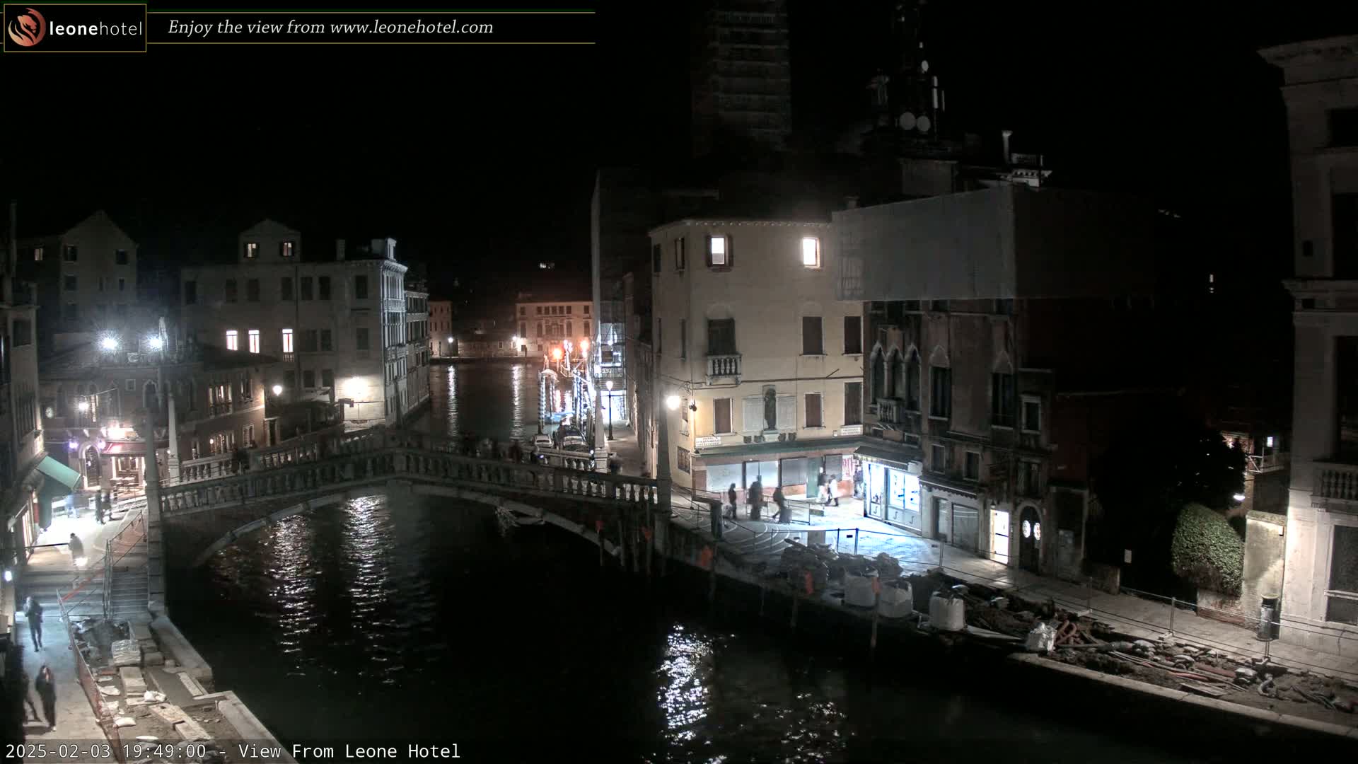 A nighttime view of a canal in Venice, Italy, showing a bridge, buildings along the waterway, and some pedestrians.