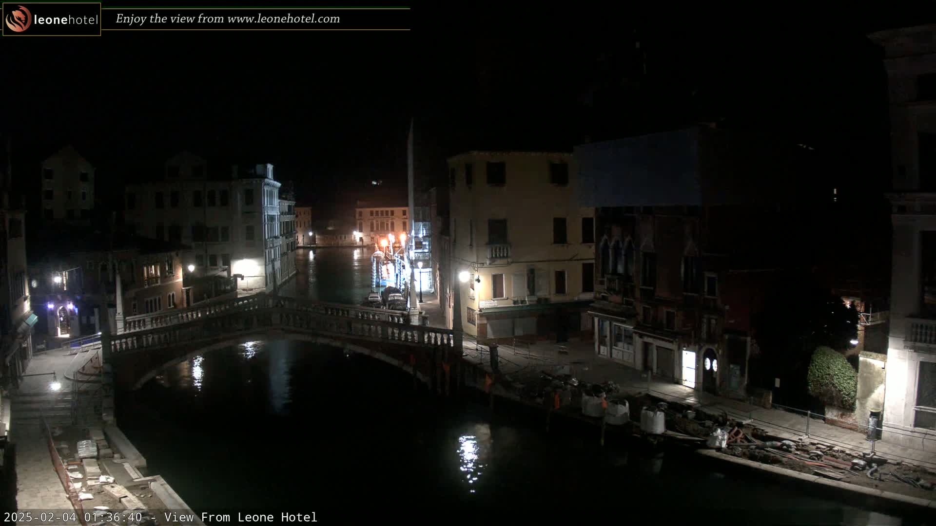 A nighttime view of a canal in Venice, Italy, shows a bridge and several buildings along the waterway, with some construction materials visible near the water's edge.