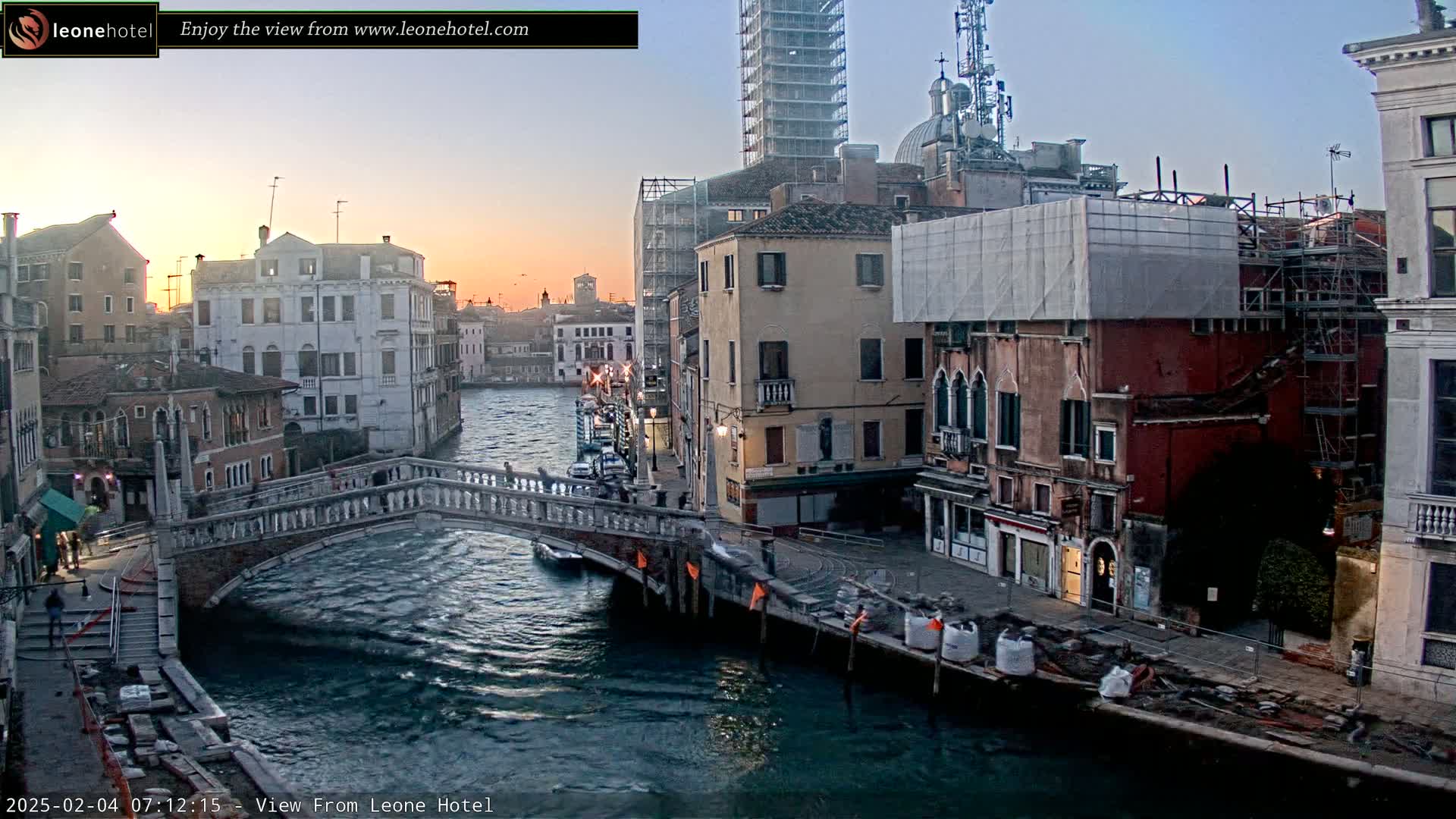 A twilight view of a Venetian canal, showing several buildings, a stone bridge, and construction materials along the water's edge.