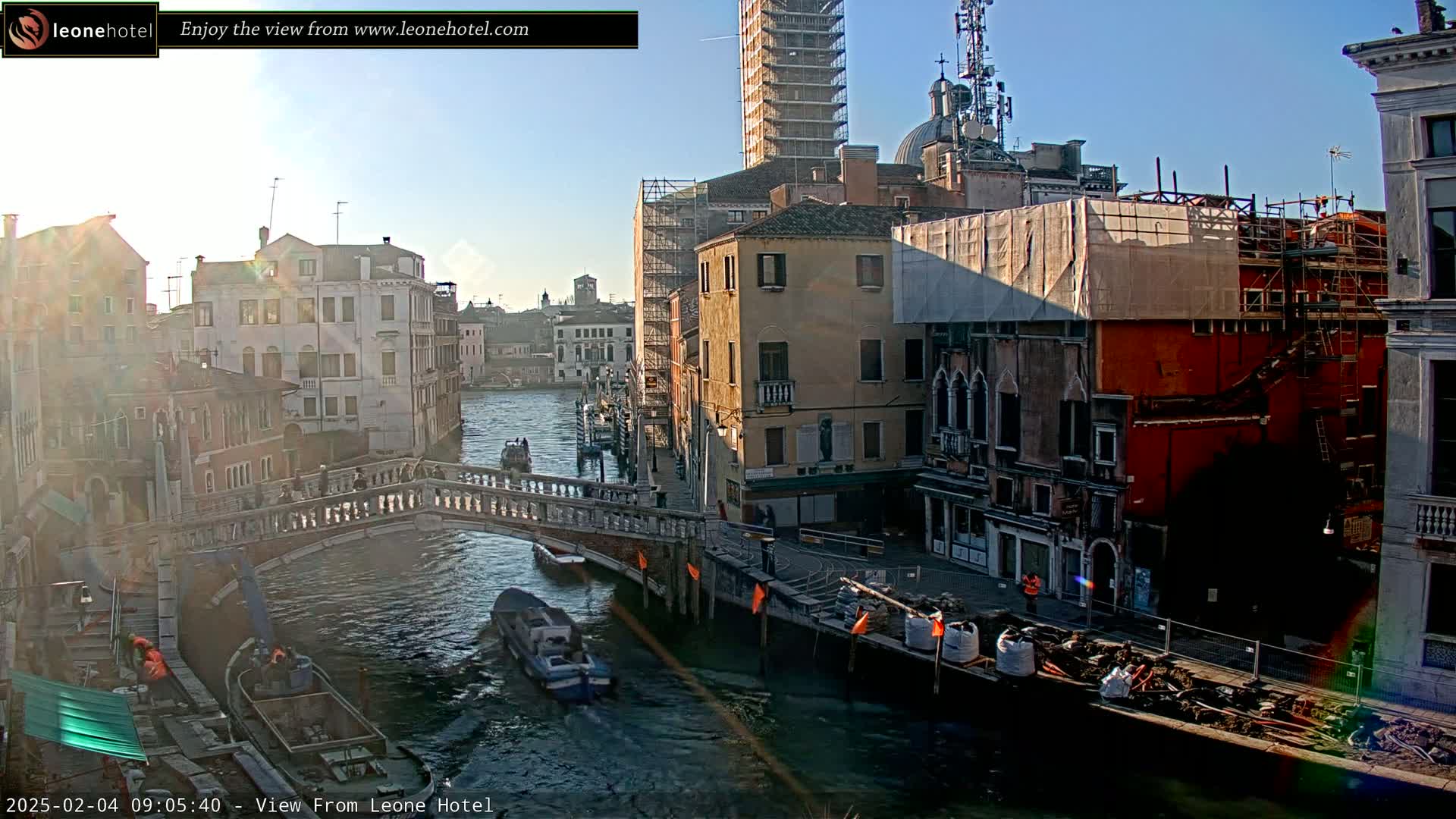 A sunny day in Venice shows a canal with boats, pedestrians on a bridge, and buildings under construction along the waterway.