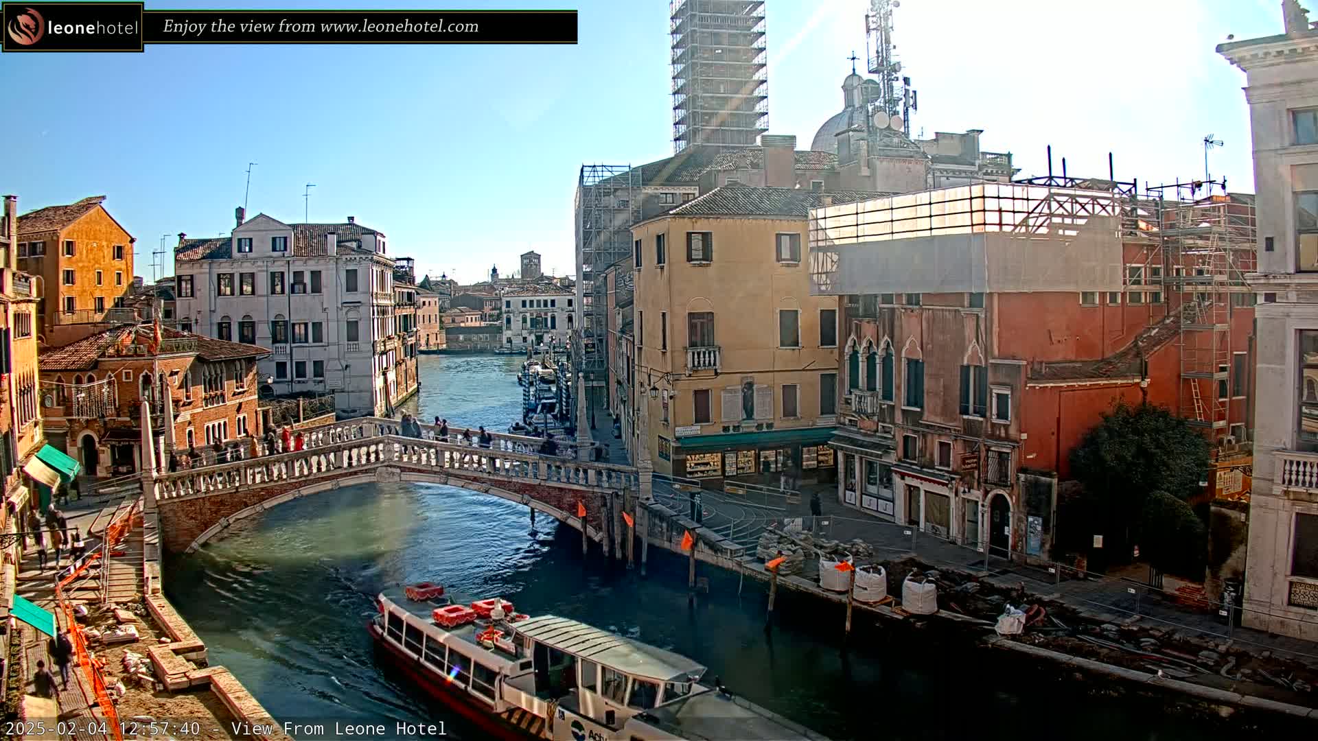 A sunny day in Venice shows a canal with a bridge, boats, and several buildings, some of which are under construction.
