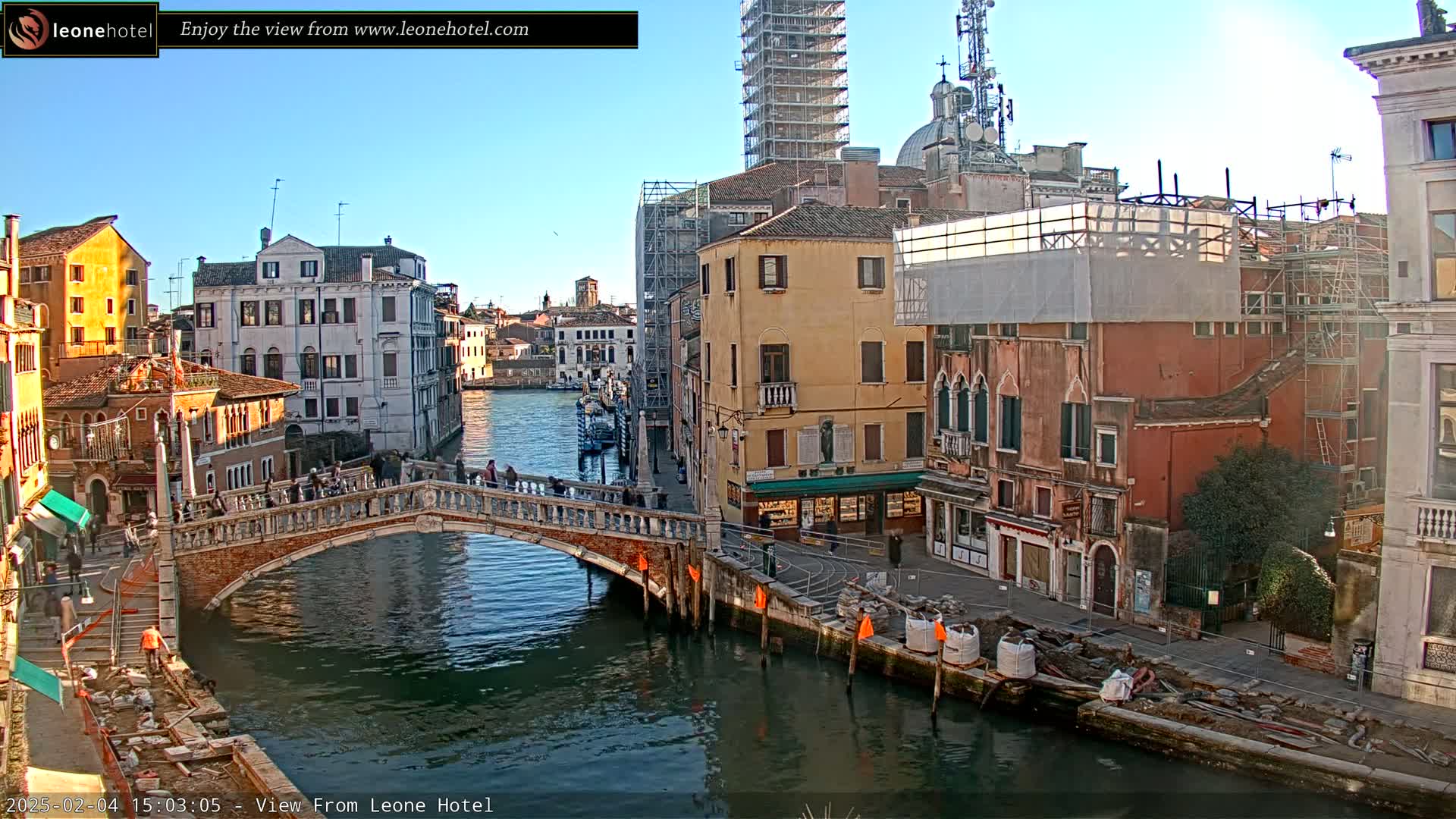 A sunny day in Venice shows a canal with a stone bridge, several buildings along the waterway, some under construction, and boats on the water.