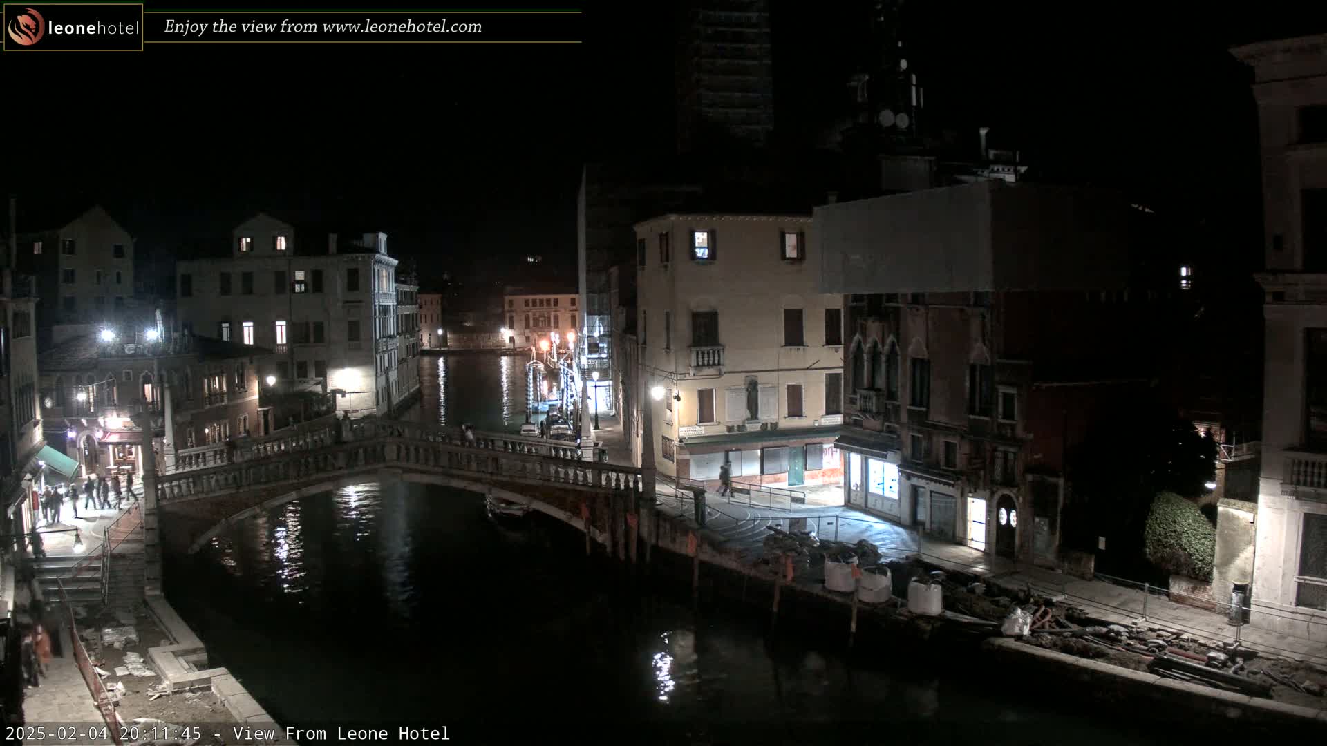 A nighttime view of a canal in Venice, Italy, shows several buildings lit along the waterway and a bridge with people crossing it.
