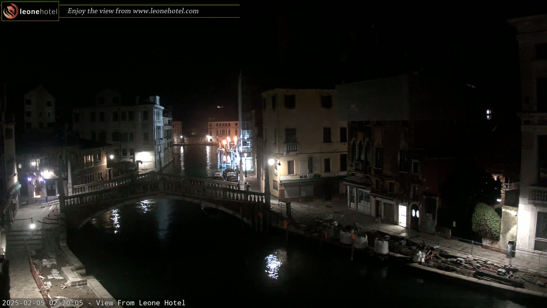 A nighttime view of a canal in Venice, Italy, shows a bridge and several buildings along the waterway, with construction visible along the canal bank.