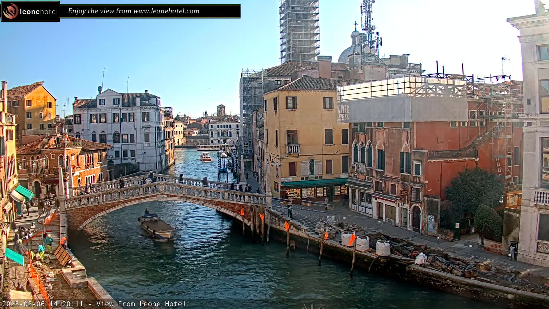 A sunny day in Venice shows a canal with a stone bridge, boats, and colorful buildings lining the waterway, some undergoing construction.