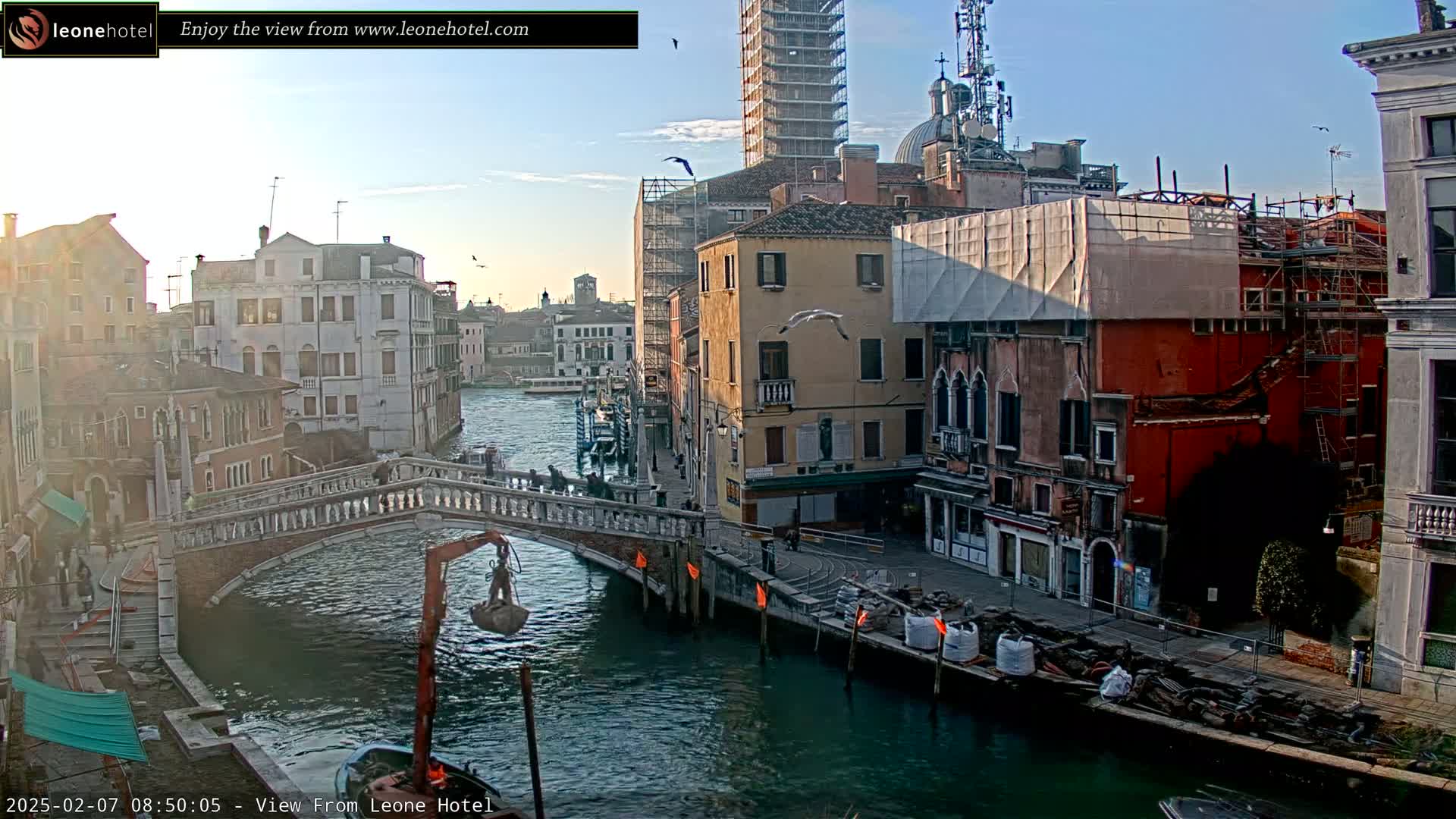 A canal in Venice, Italy, under a mostly clear sky at sunrise, shows several buildings, a bridge, and construction activity along the waterway.