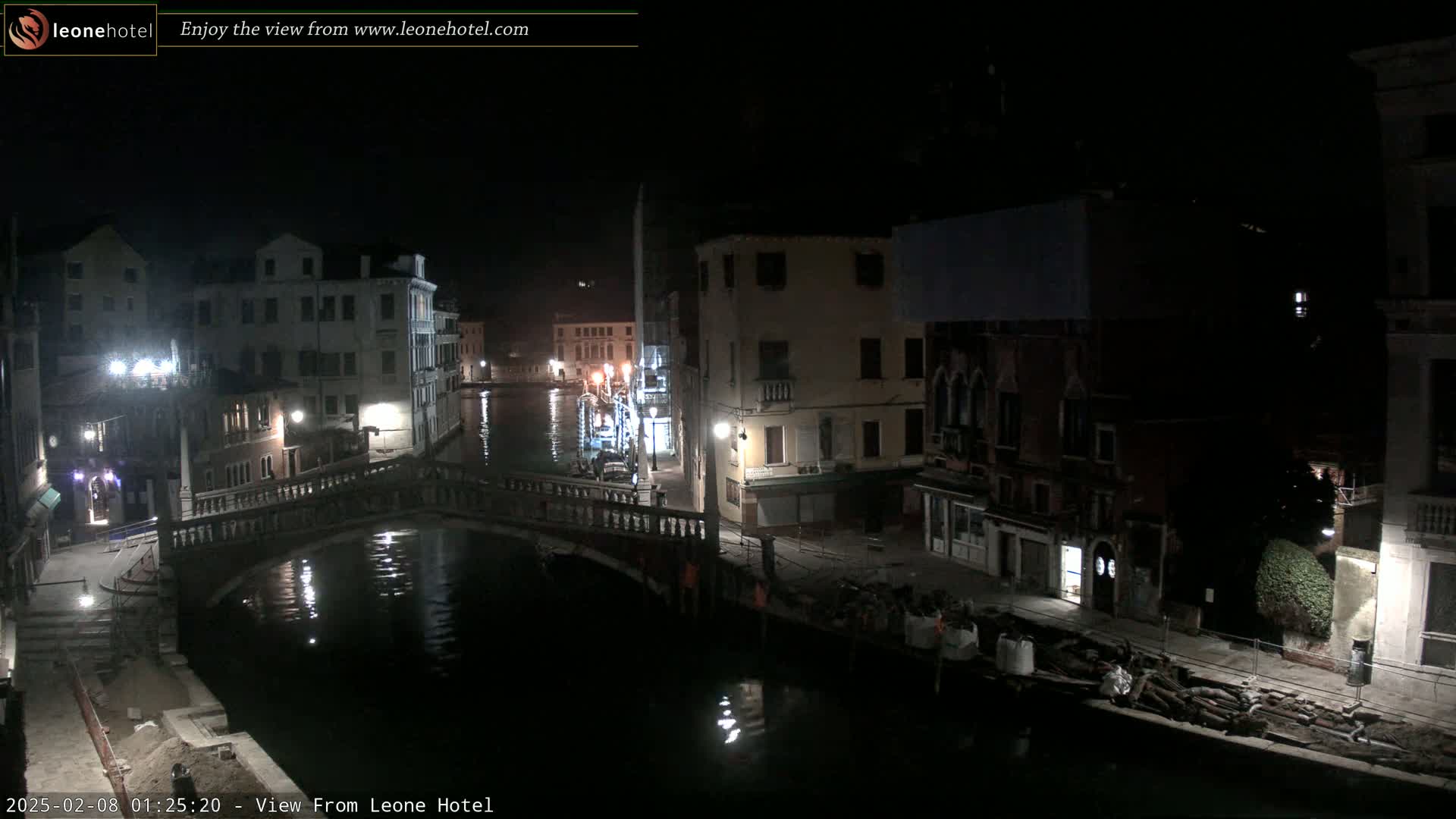 A nighttime view of a canal in Venice, Italy, shows several buildings lining the waterway, a bridge spanning the canal, and some construction materials along the shore.