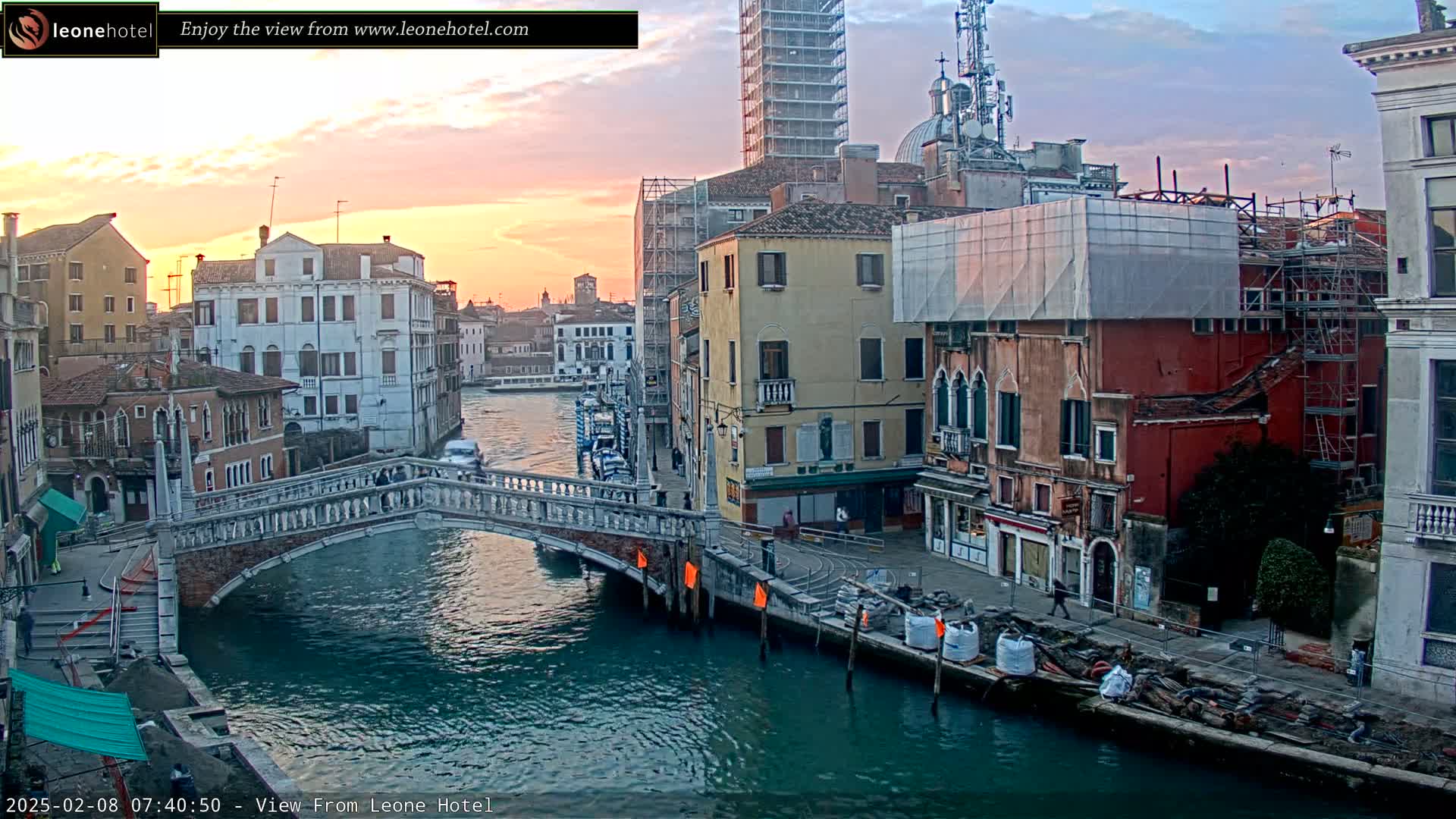 A sunset illuminates a canal in Venice, Italy, where several stone bridges connect buildings undergoing renovations.