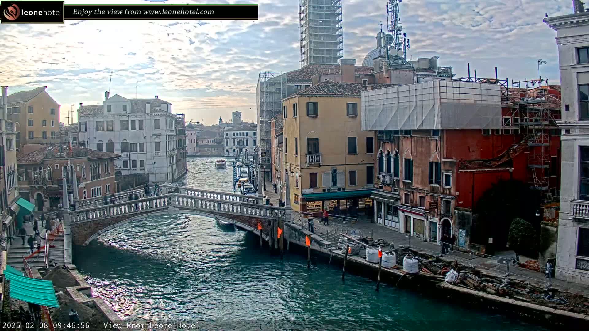 A canal in Venice, Italy, under a partly cloudy sky, shows a stone bridge, several buildings, some under construction, and boats on the water.