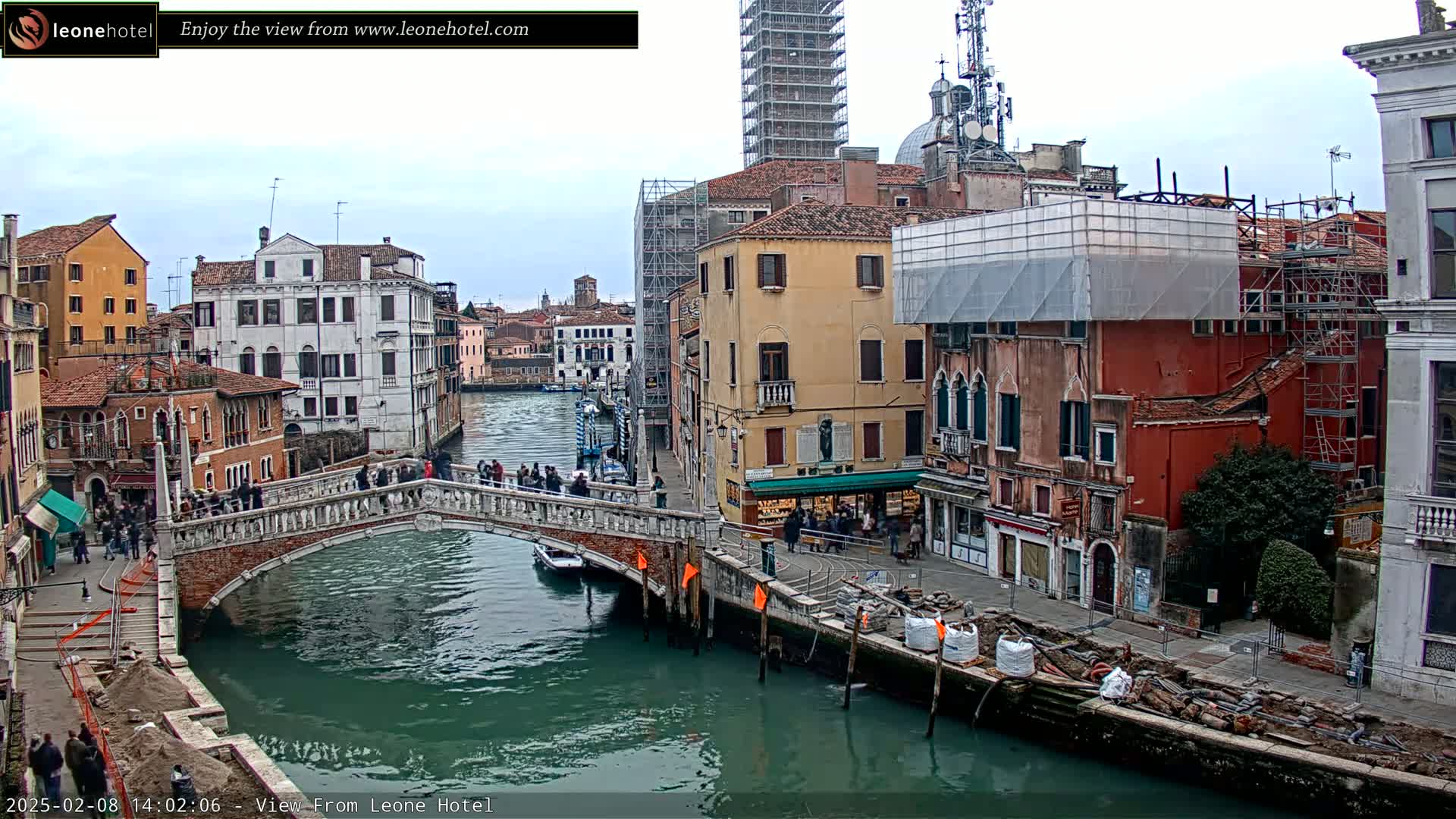 A canal in Venice, Italy, with overcast skies, shows a stone bridge with people on it, colorful buildings lining the waterway, and construction scaffolding visible on some structures.