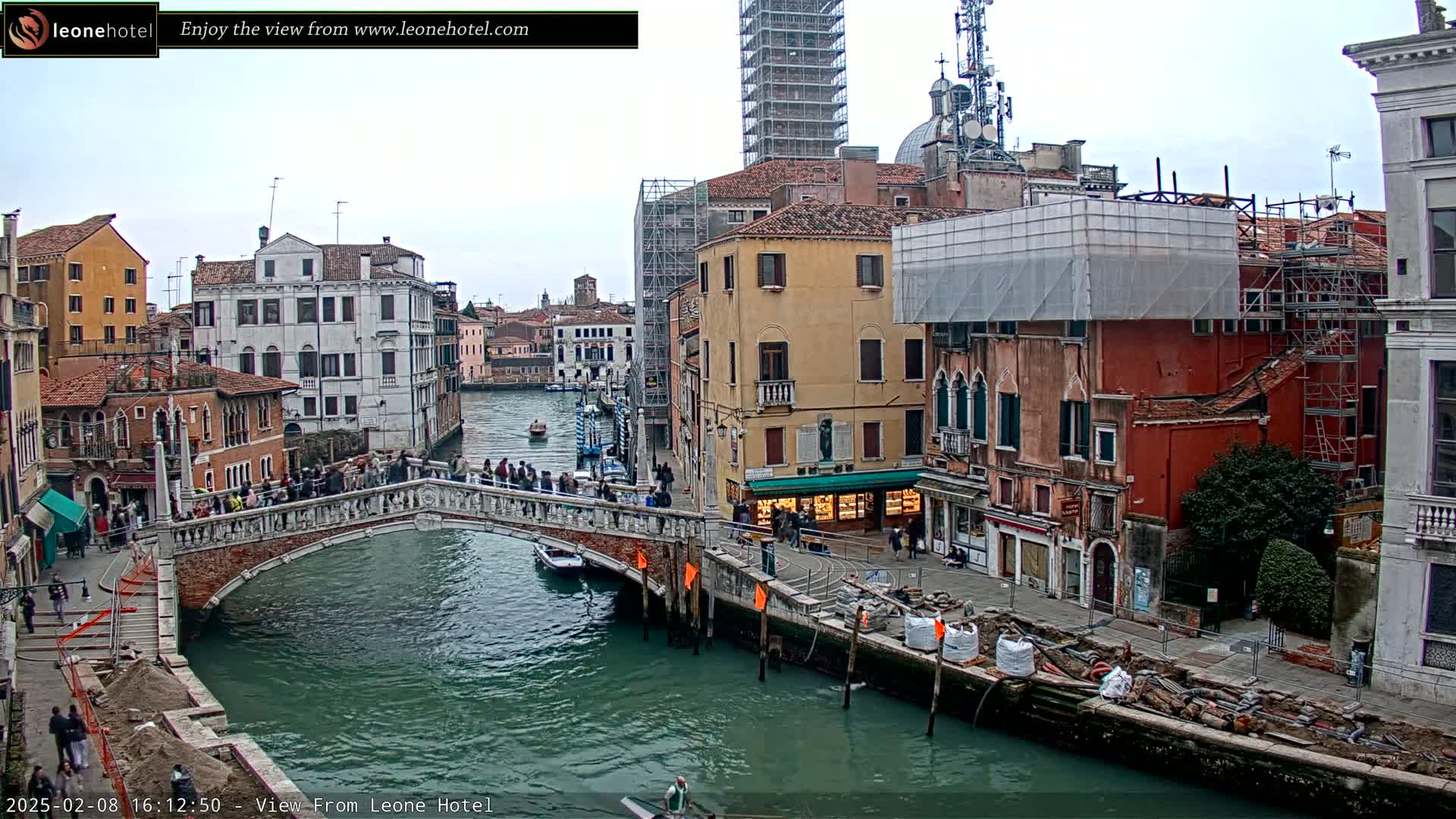 A bridge over a canal in Venice, Italy, is crowded with people under an overcast sky, with buildings under construction visible along the waterway.