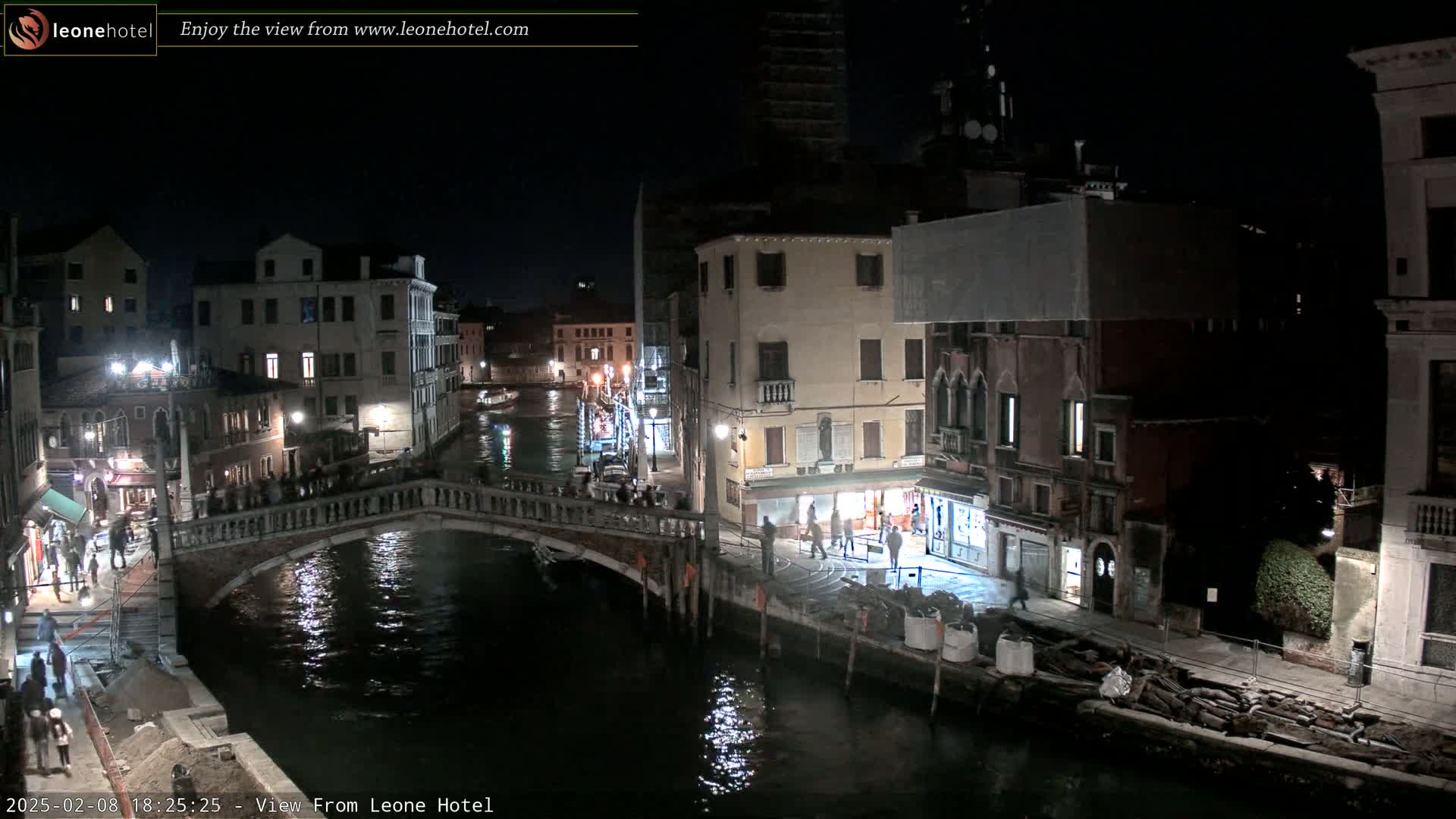 A nighttime view of a canal in Venice, Italy, shows a stone bridge over the water with buildings lining both sides, and people walking along the canal.