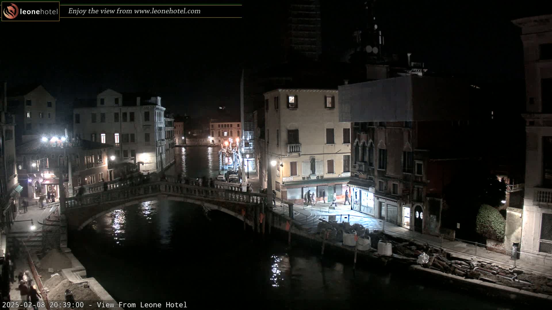 A nighttime view of a canal in Venice, with bridges, buildings, and a few people visible under a dark sky.