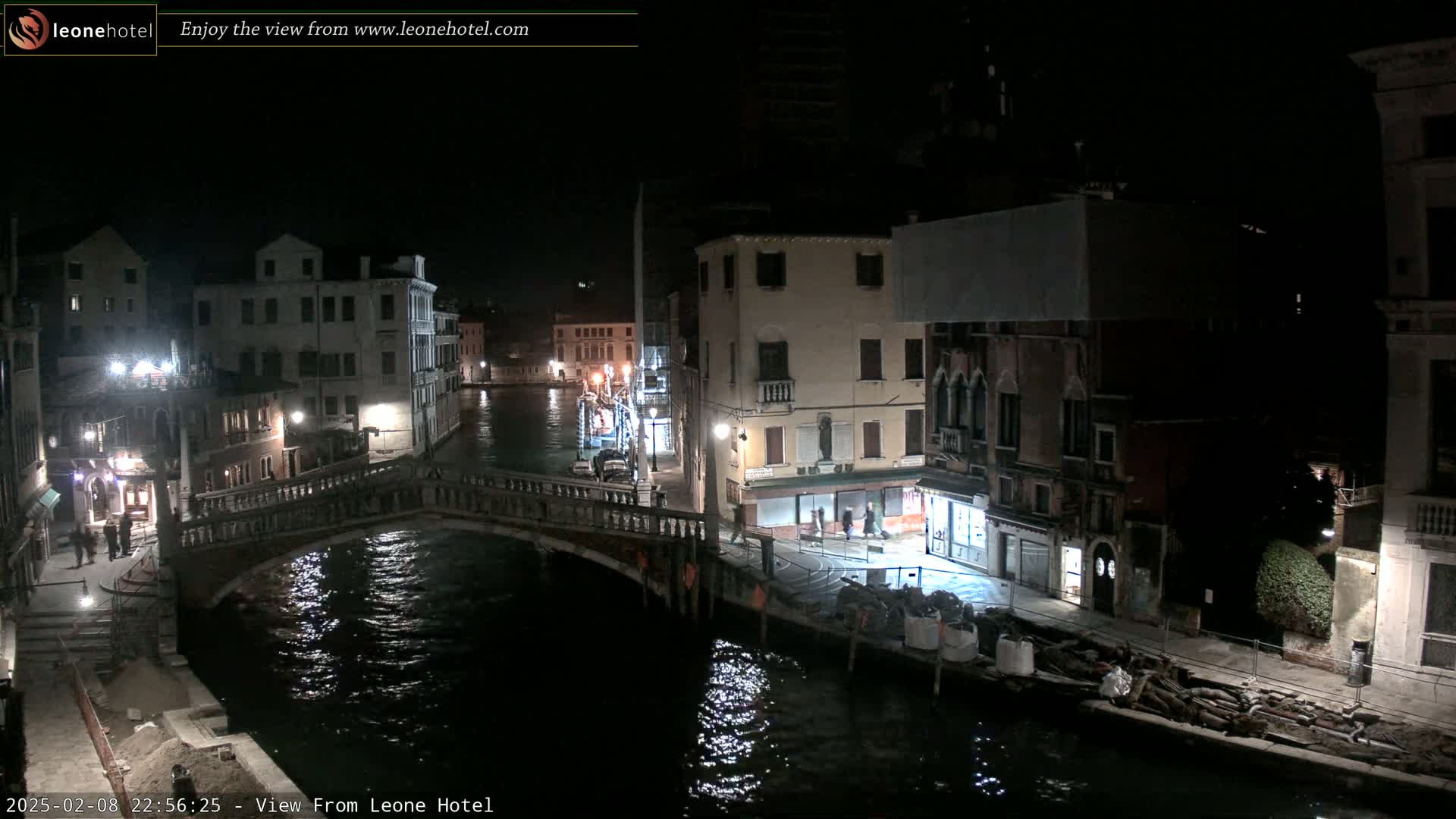 A nighttime view of a canal in Venice, Italy, showing bridges, buildings, and a few people walking along the water's edge.