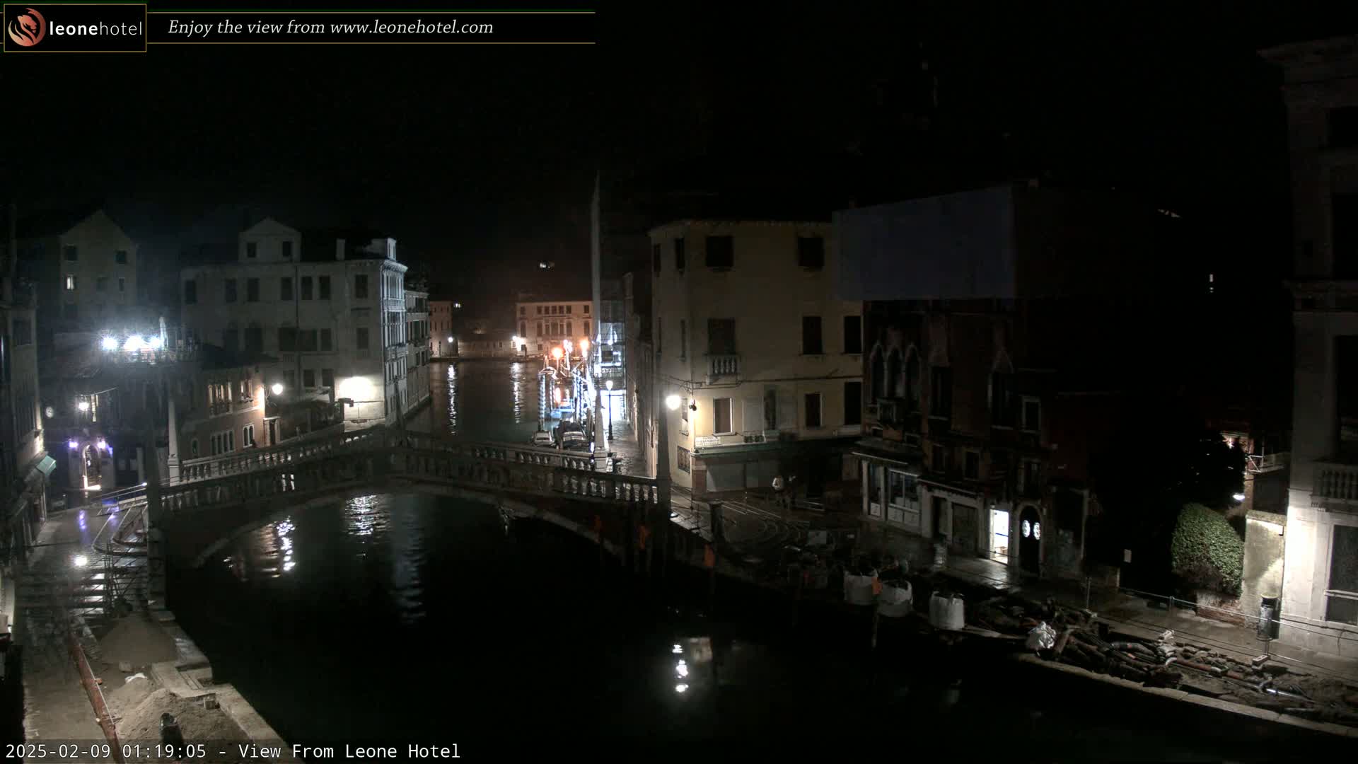 A nighttime view of a canal in Venice, showing several buildings along its banks and a bridge crossing the waterway, with wet pavement and construction materials visible near the water's edge.
