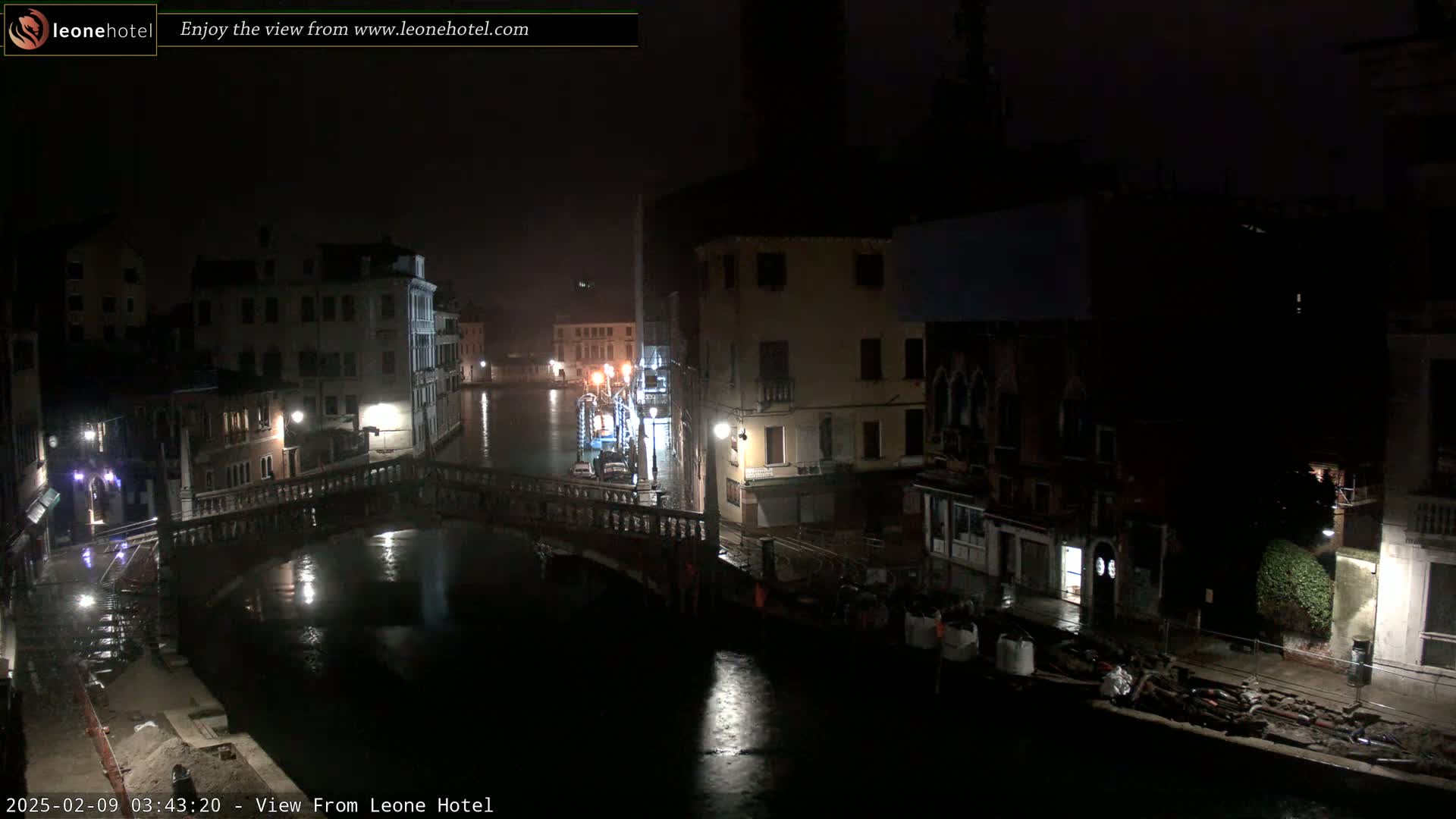 A nighttime view of a canal in Venice, Italy, shows a bridge and buildings along the waterway under dark, possibly rainy conditions.