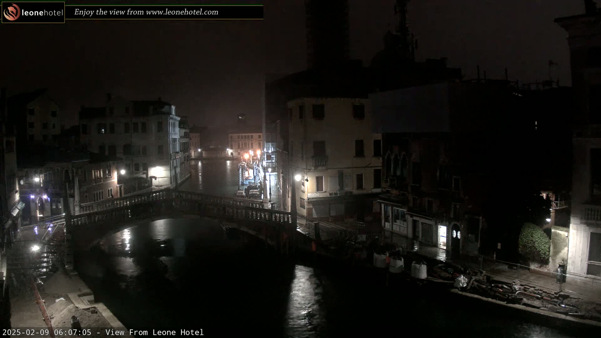 A nighttime view of a canal in Venice, showing several buildings lining the waterway and a bridge crossing the canal under rainy conditions.