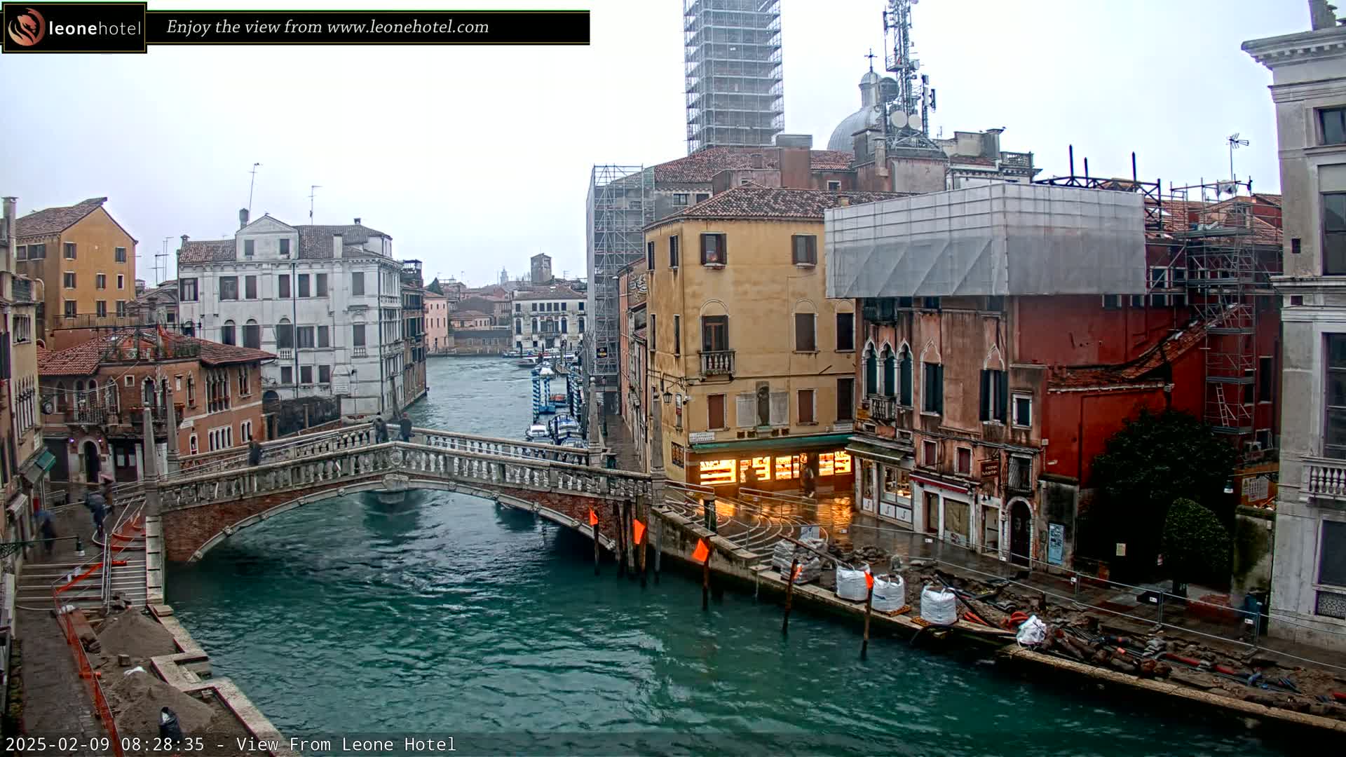 A view of a canal in Venice, Italy, on an overcast day, shows a stone bridge, several buildings under construction, and boats on the water.