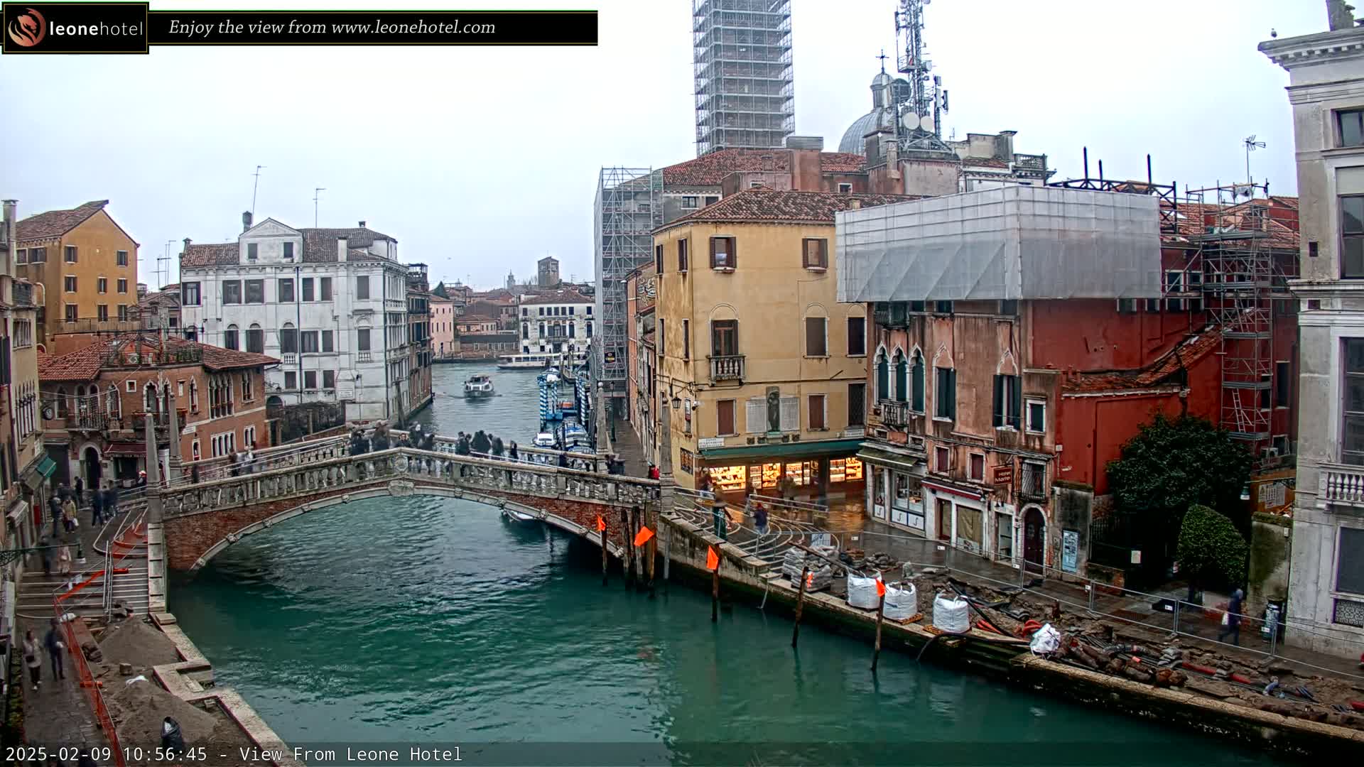 A view of a canal in Venice, Italy, on an overcast day, shows several buildings, a bridge with people crossing it, and some construction materials along the water's edge.