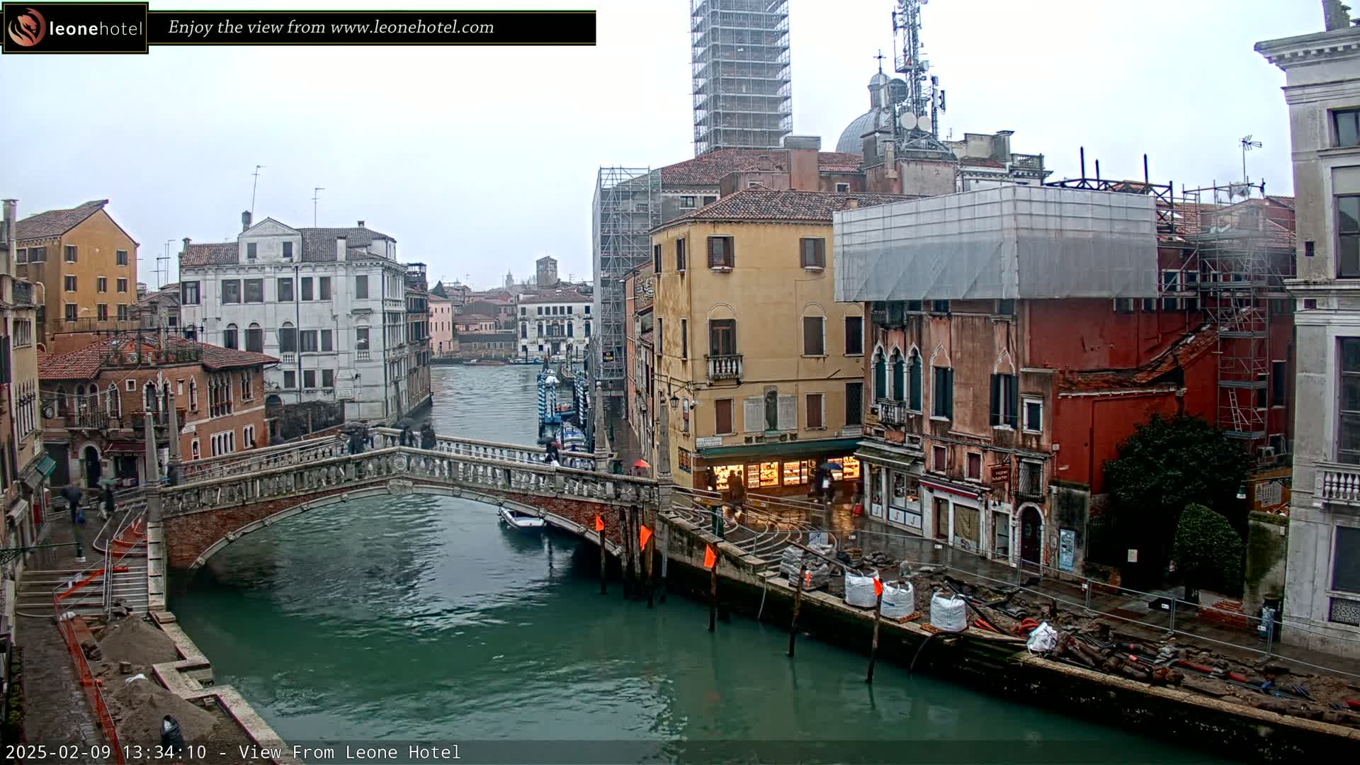 A view of a canal in Venice, Italy, on an overcast day, shows a stone bridge with pedestrians, colorful buildings along the waterway, and construction materials on the canal bank.