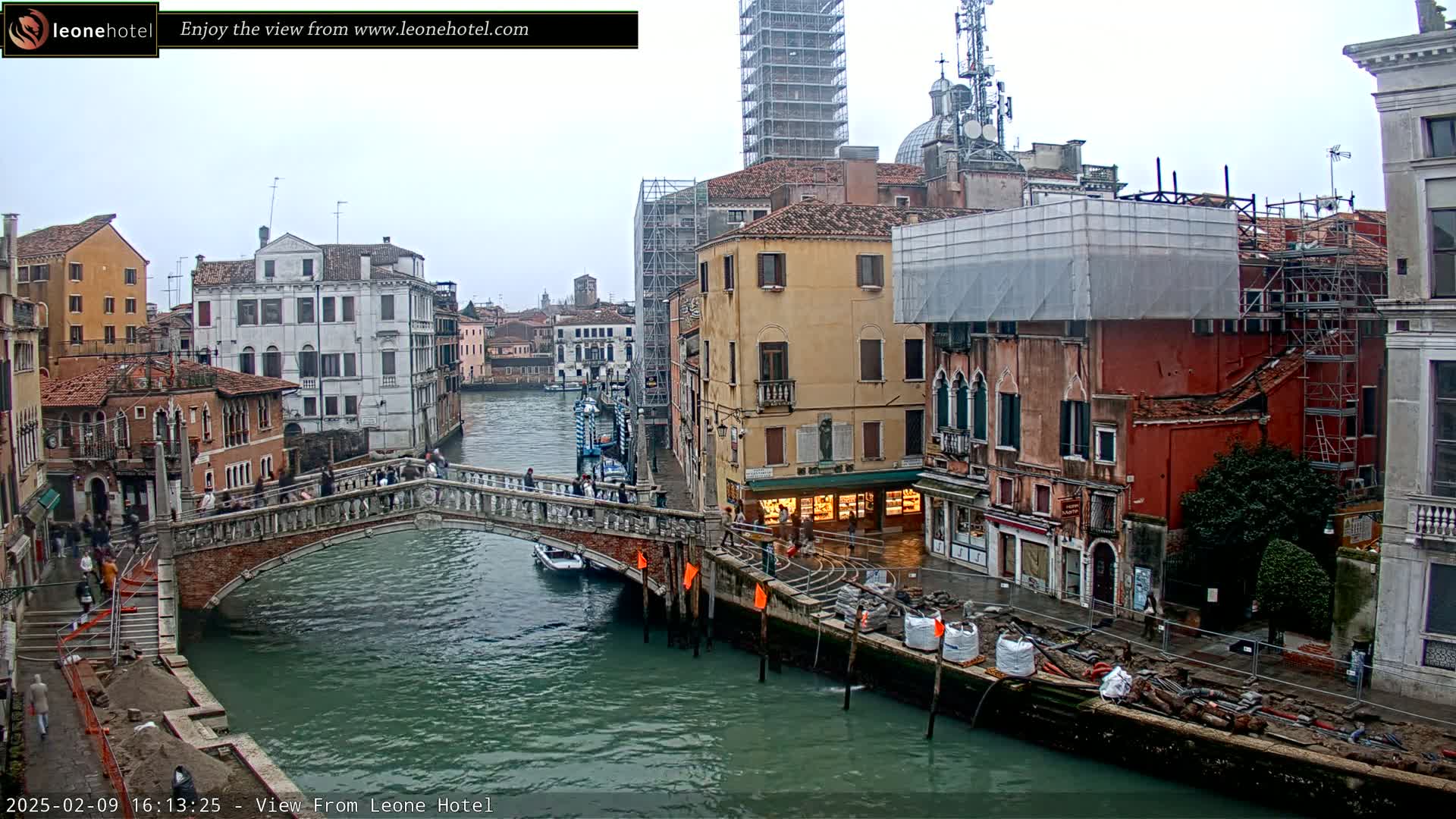 A canal in Venice, Italy, on an overcast day, shows a stone bridge with people crossing it, and buildings along the waterway under construction, with scaffolding and building materials visible on the canal bank.