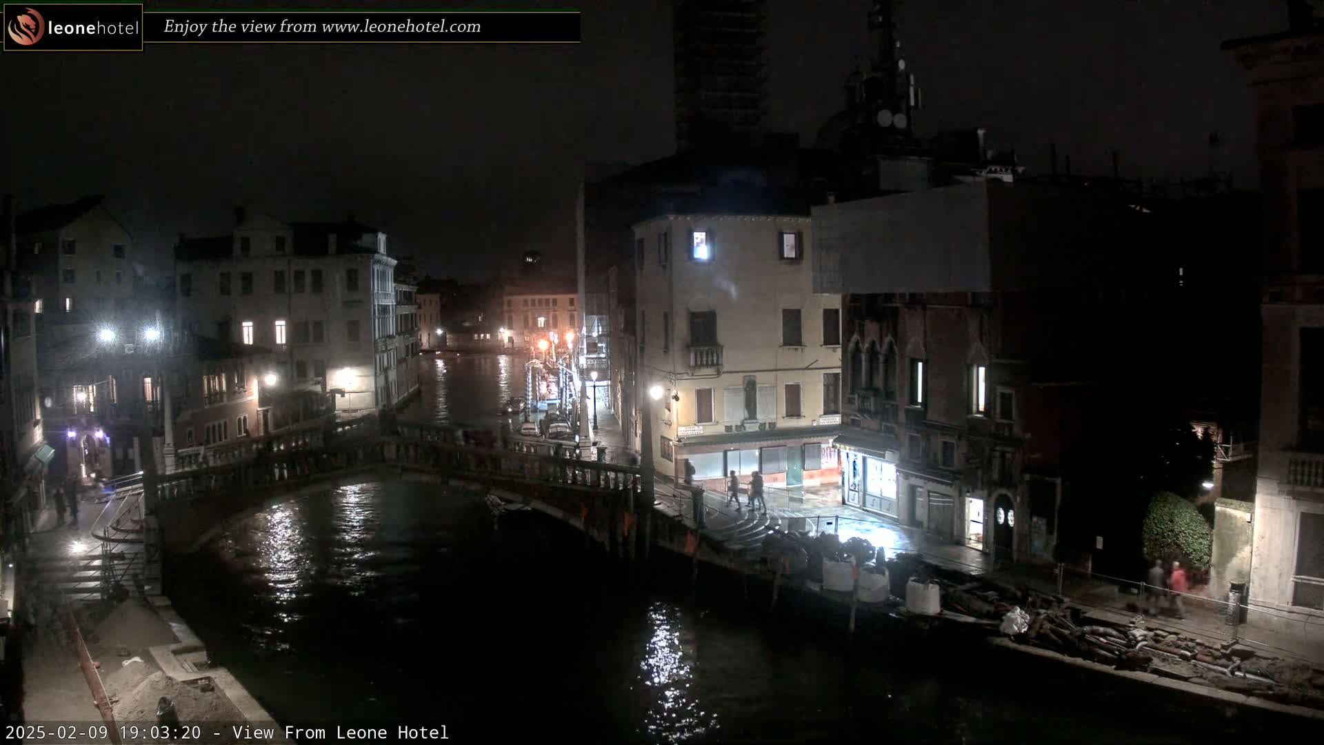 A nighttime view of a canal in Venice, Italy, shows several buildings illuminated along the water's edge, with a bridge crossing the canal and some people walking along the banks.