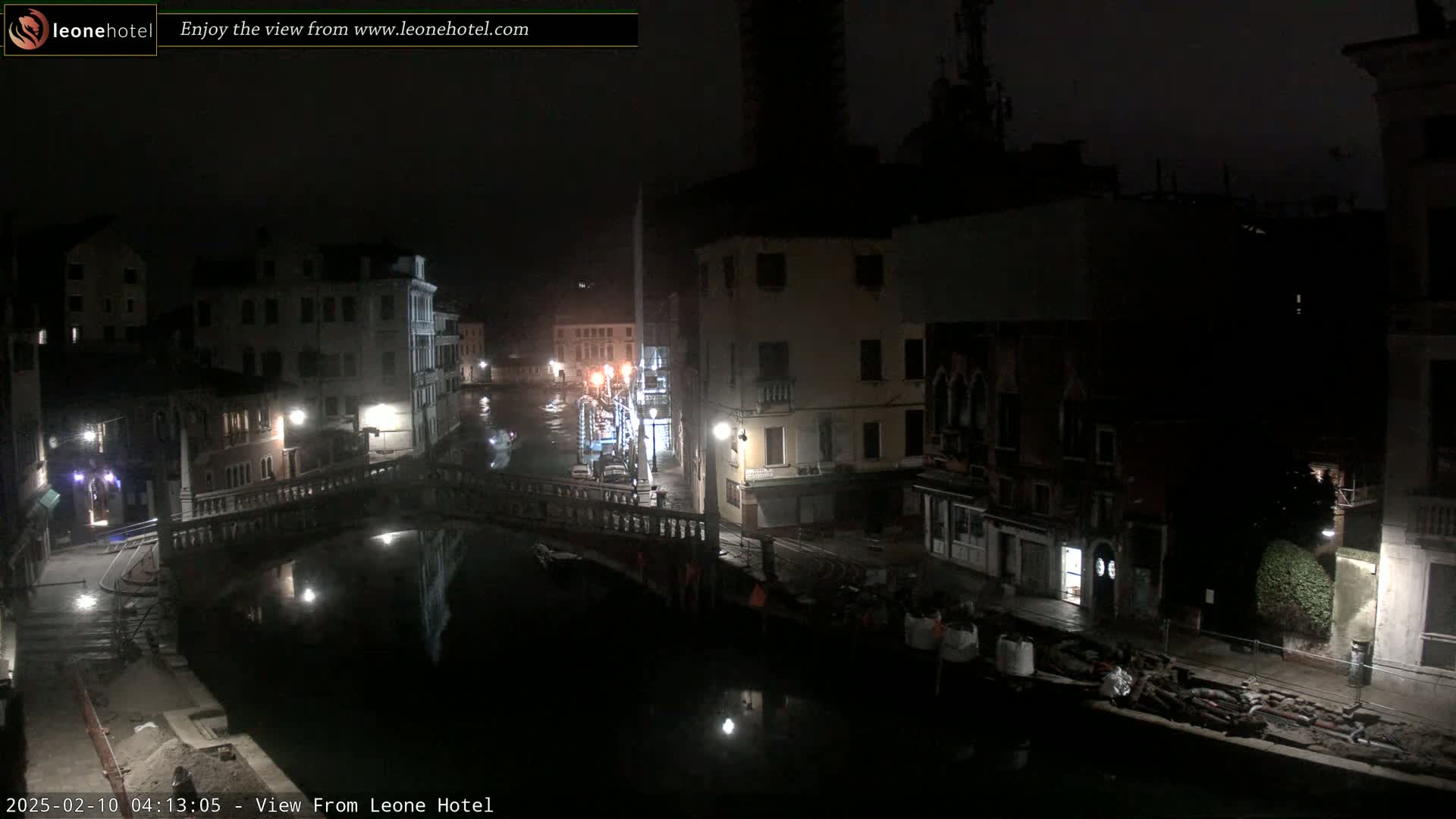A nighttime view of a canal in Venice, Italy, shows several buildings lining the waterway, with a bridge crossing the canal and some activity visible along the water's edge.