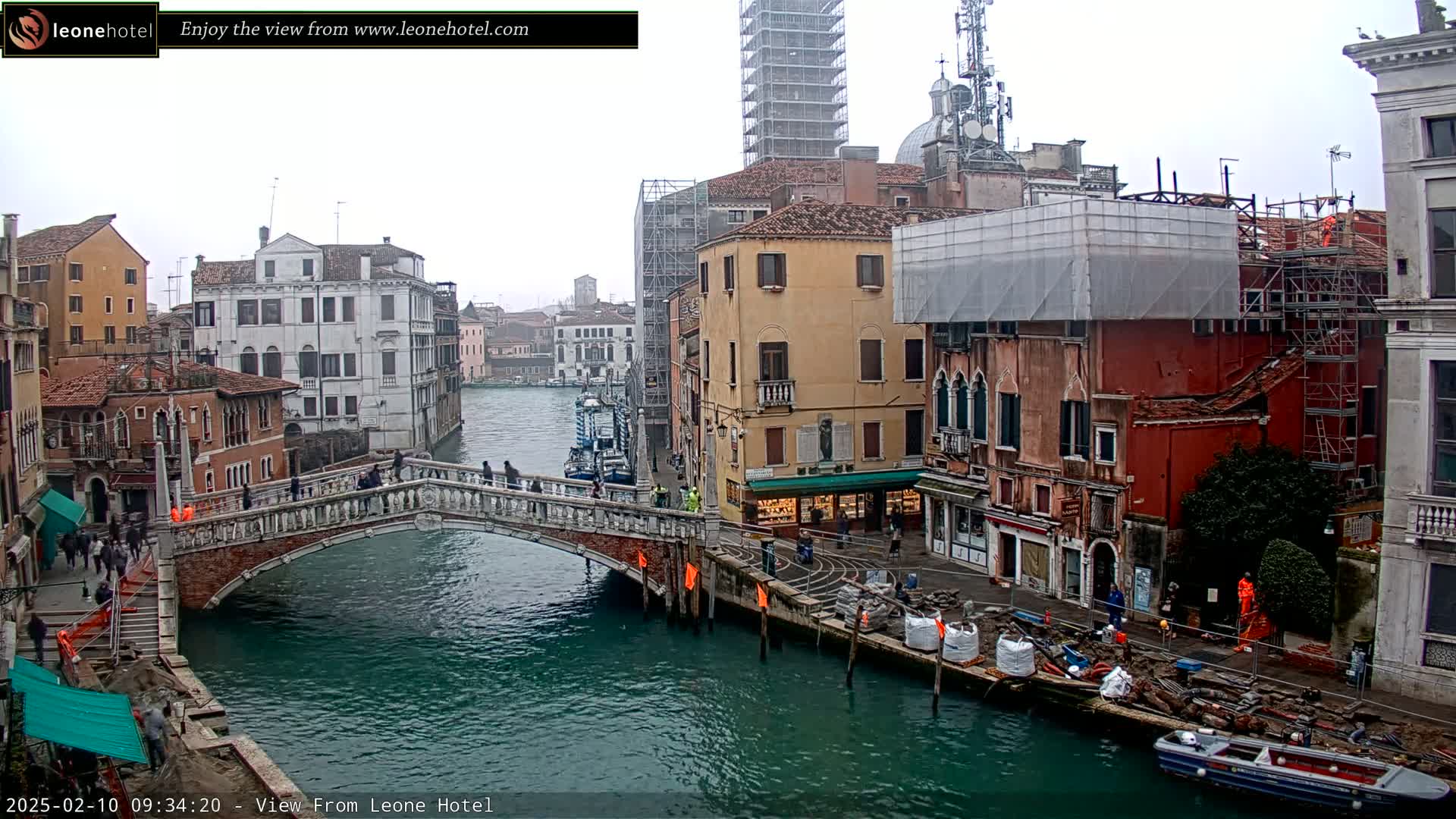 A view of a canal in Venice, Italy, on an overcast day, shows a stone bridge with pedestrians, buildings under construction, and a boat on the water.