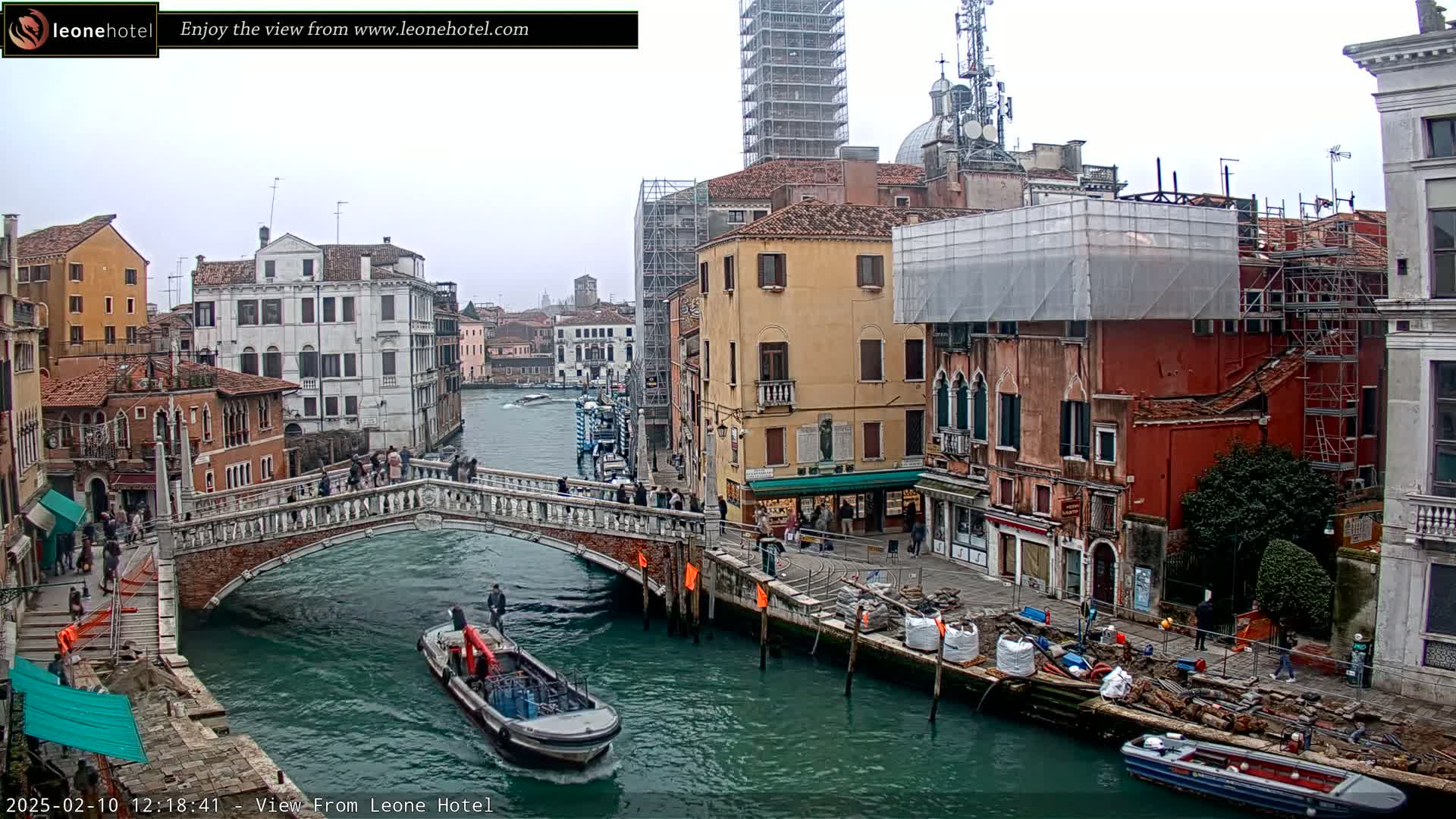 A view of a Venetian canal on an overcast day, showing a stone bridge, boats, and buildings under construction.