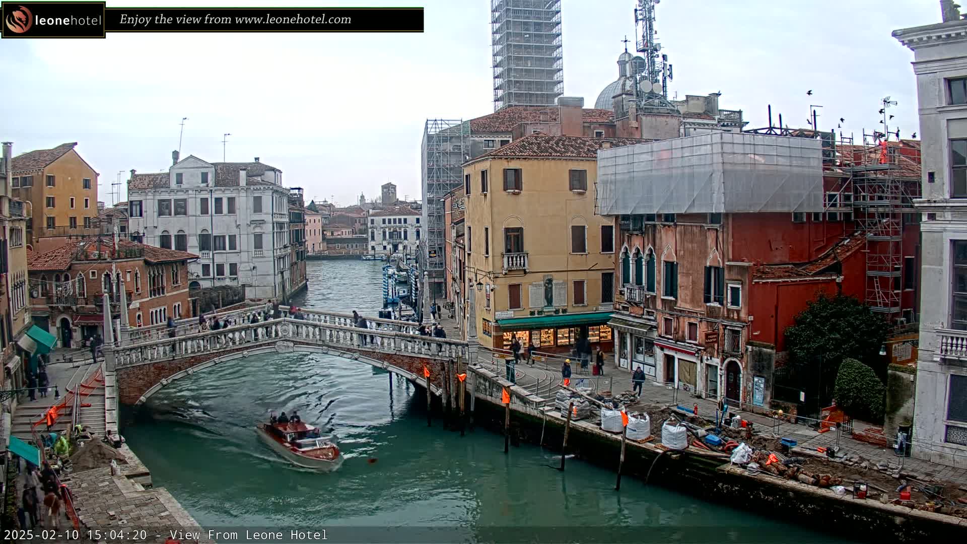 A Venetian canal scene on an overcast day shows a stone bridge, several buildings under construction, a motorboat on the water, and construction materials along the canal.