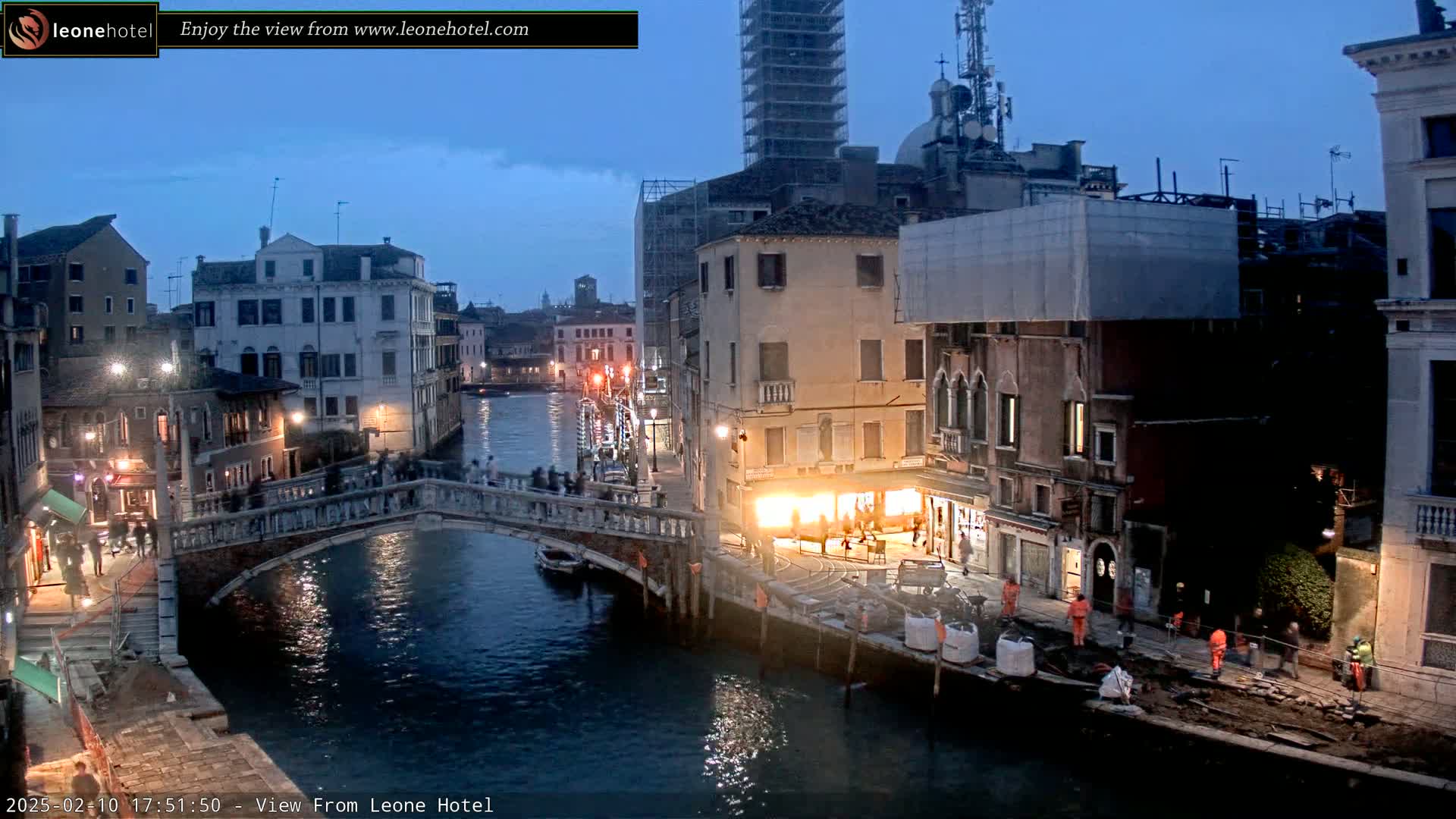 A twilight view of a Venetian canal, showing a bridge with people crossing, buildings lining the waterway, and construction activity near the water's edge.