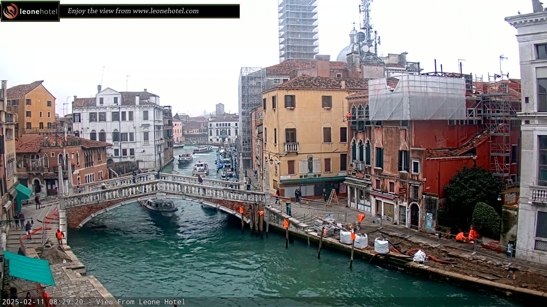 A canal in Venice, Italy, on an overcast day, shows several buildings, a bridge, boats, and construction along the waterway.
