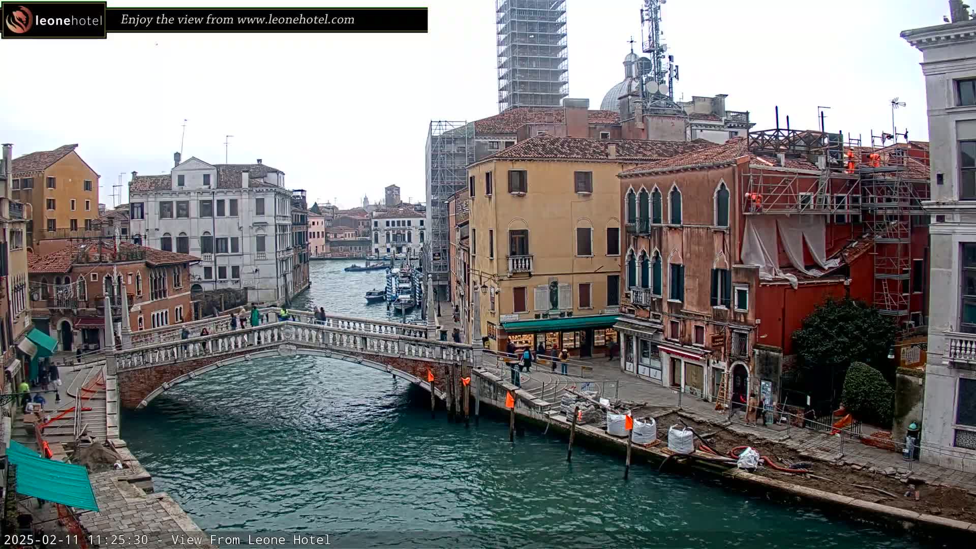 A bridge crosses a canal in Venice, Italy, under an overcast sky; several buildings line the canal, one of which is under construction.