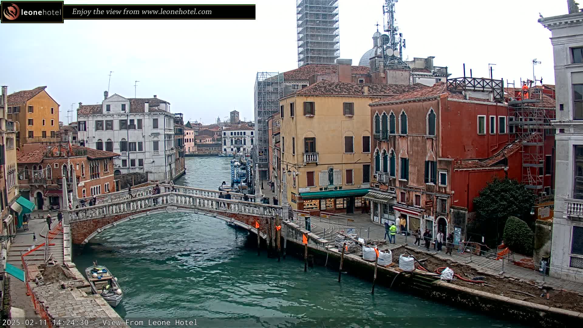 A stone bridge arches over a canal in Venice, Italy, under an overcast sky, with colorful buildings lining the waterway and construction underway near the canal's edge.