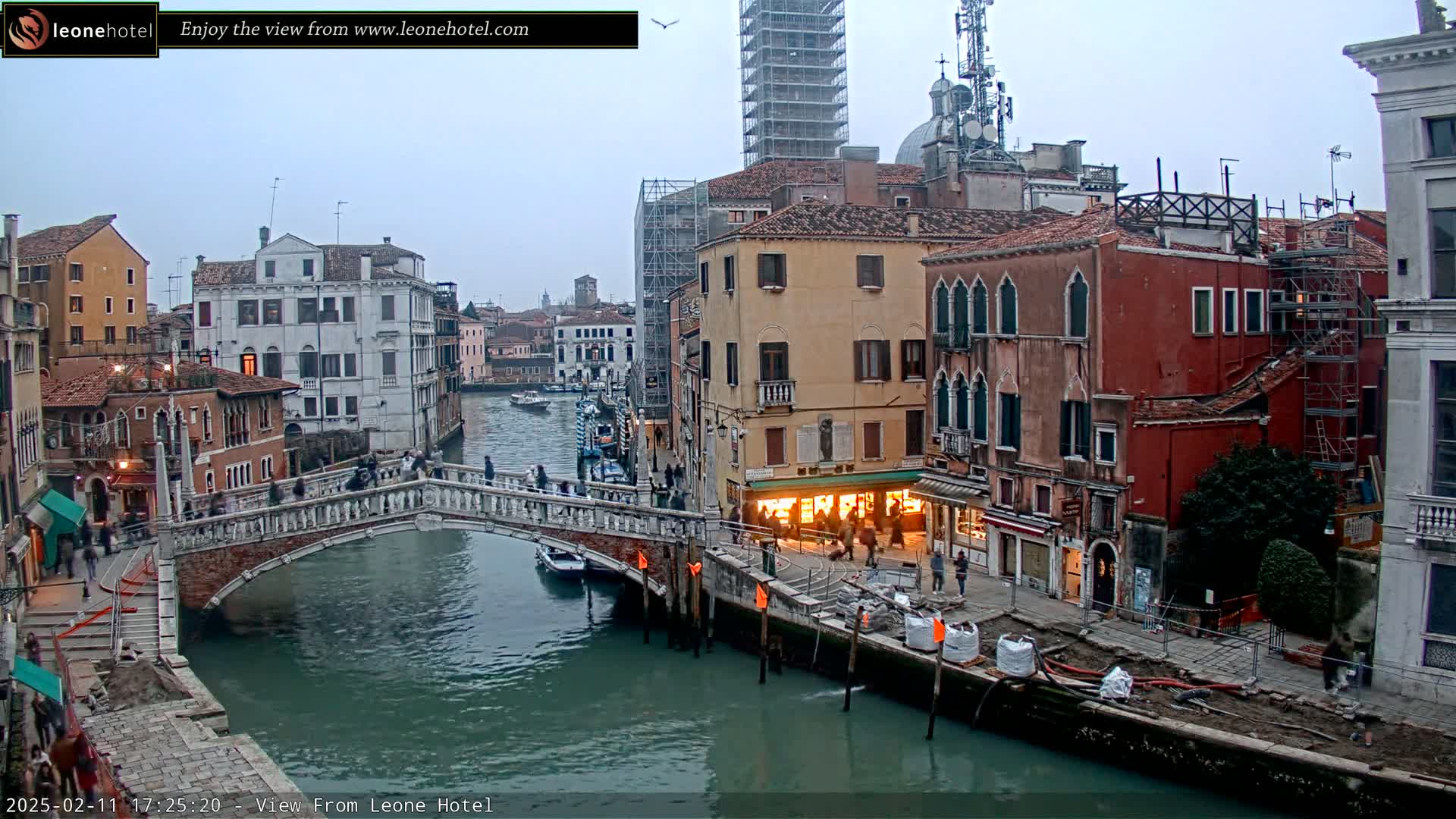 A stone bridge spans a canal in Venice, Italy, under an overcast sky, with several buildings lining the waterway, some under construction.