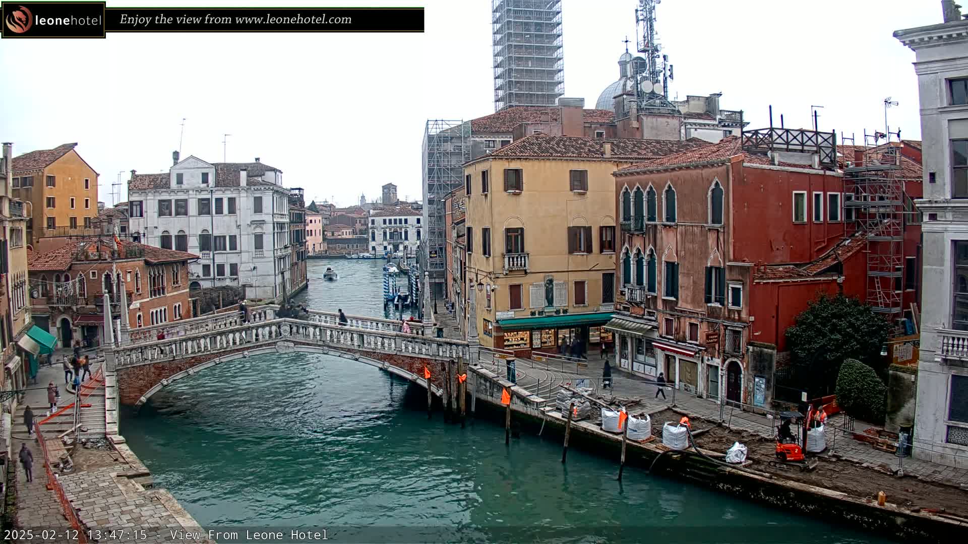 A Venetian canal scene on an overcast day shows a stone bridge arching over the water, with colorful buildings lining the canal banks, some undergoing construction or renovation.