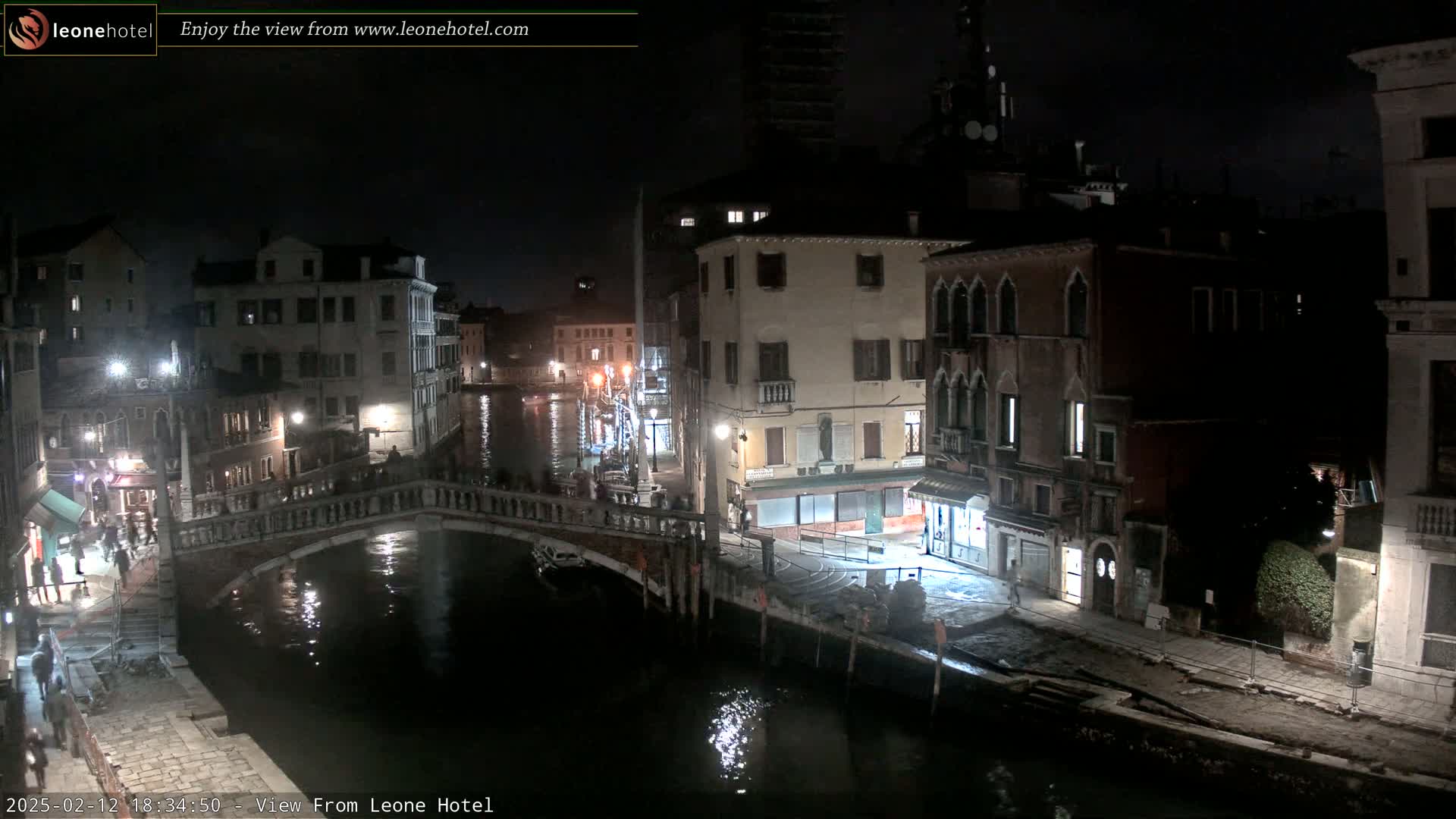 A nighttime view of a canal in Venice, Italy, shows a stone bridge over the water, with buildings lining both sides of the canal, lit by streetlights.