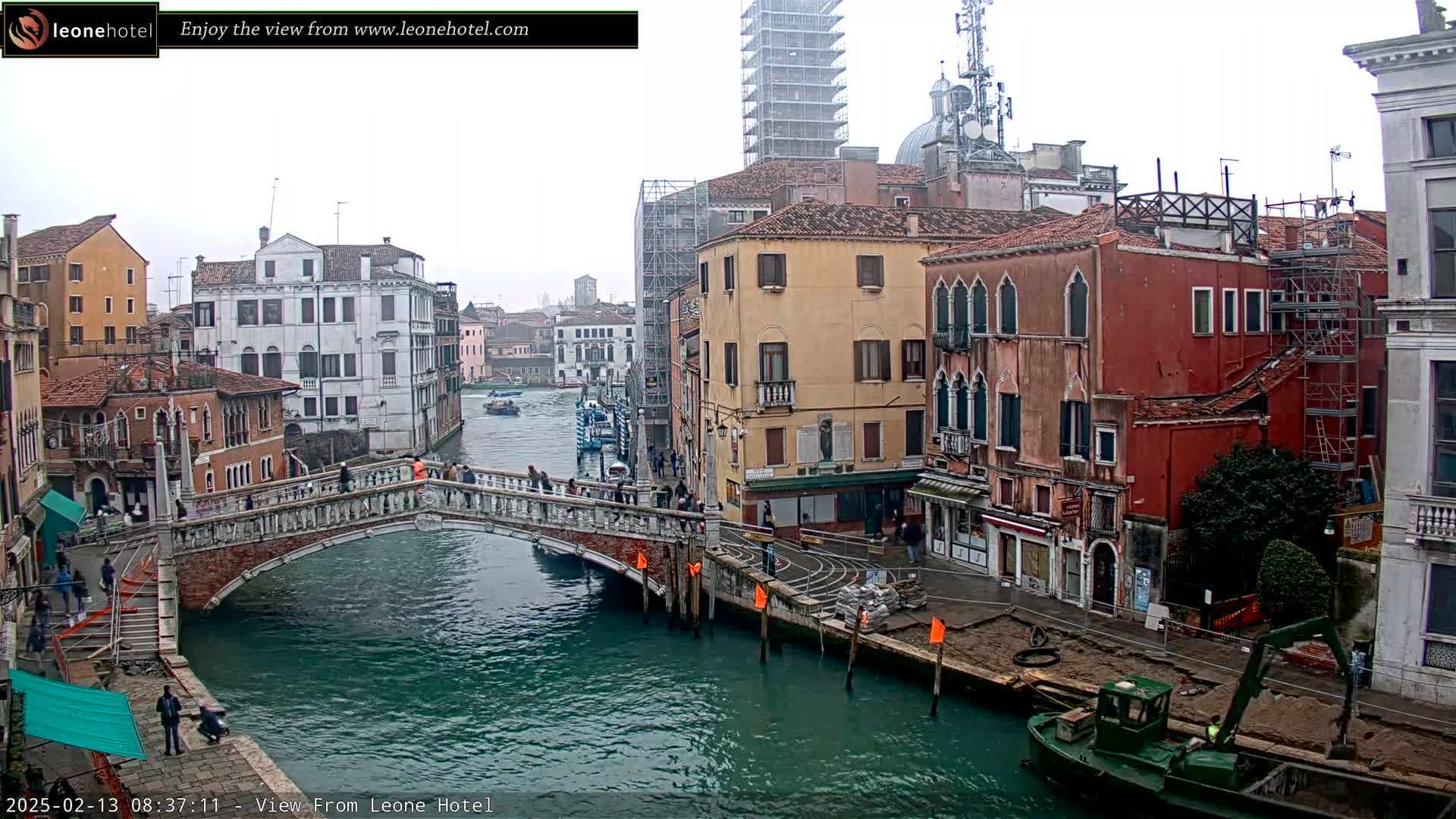 A view of a canal in Venice, Italy, on an overcast day, shows a stone bridge with people walking across it and colorful buildings lining the waterway, with construction visible on some structures and a small boat in the canal.