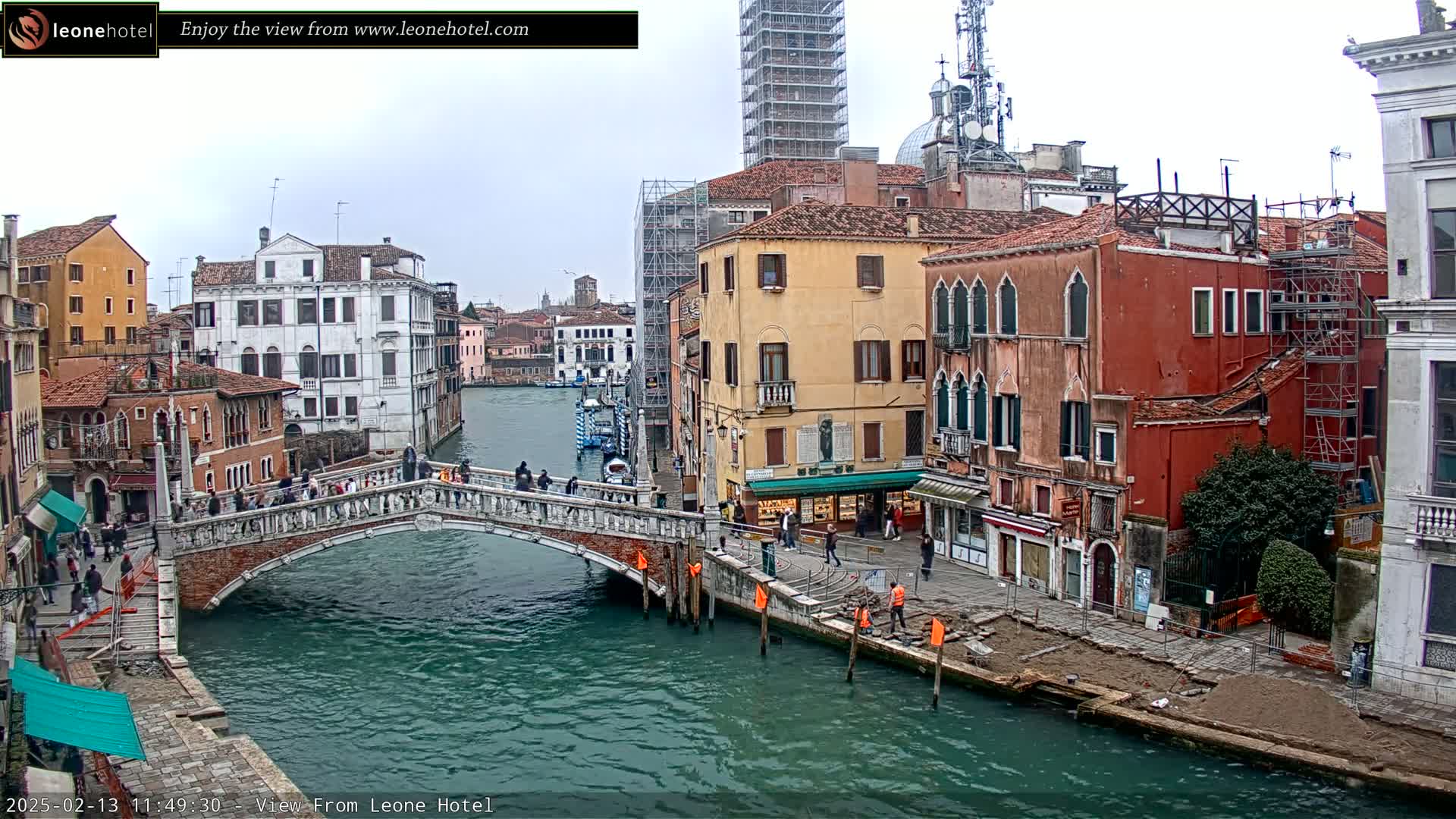 A view of a canal in Venice, Italy, on an overcast day, shows a stone bridge with people walking across it, and colorful buildings lining the waterway, some of which are under construction.