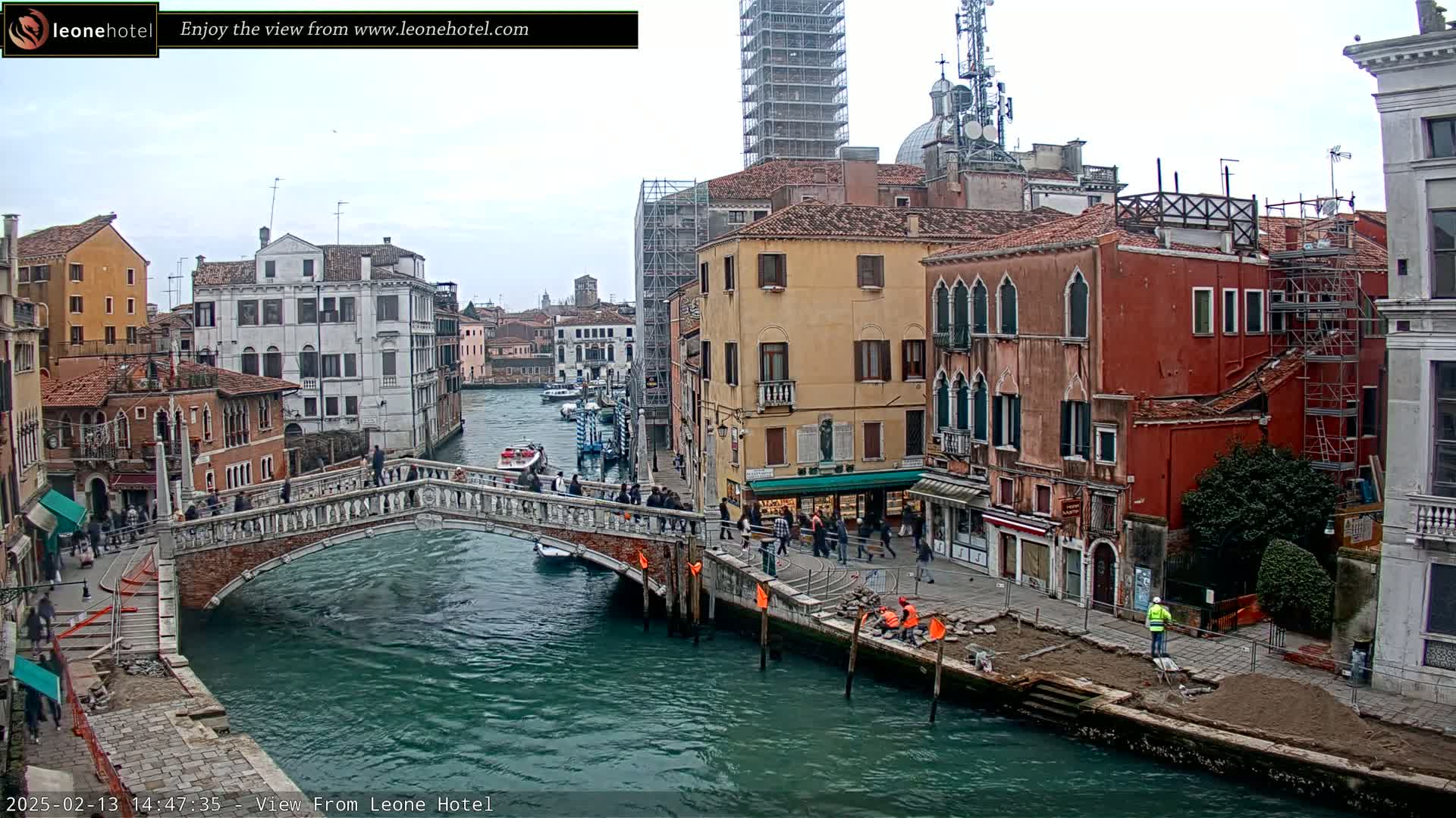 A canal in Venice, Italy, with overcast skies, shows a stone bridge over the water, lined with buildings, some under construction, and people walking along the canal.